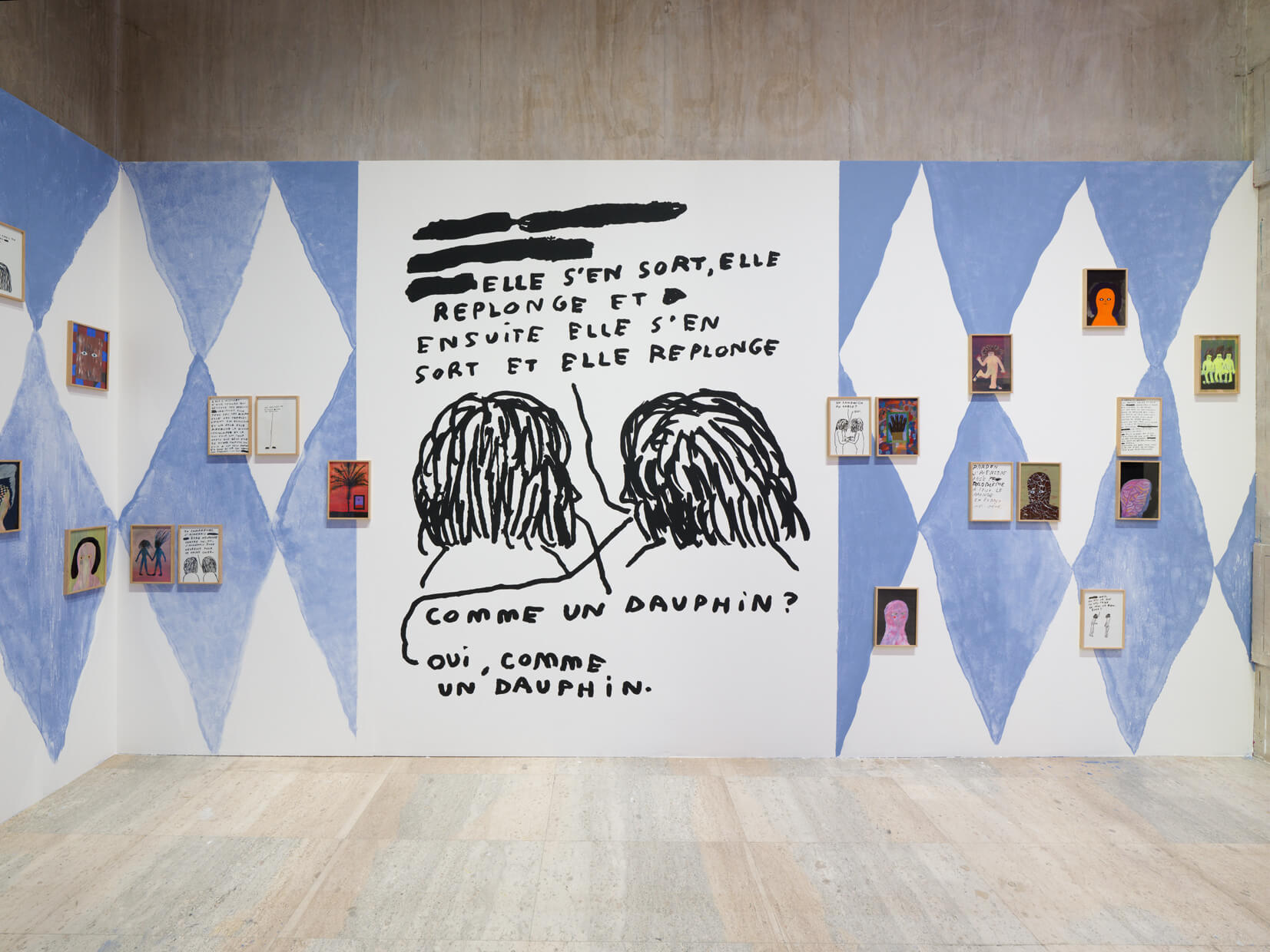 Installation artistique avec des murs à motifs de losanges bleus et blancs présentant des œuvres d'art encadrées et une grande peinture murale en noir et blanc avec un texte en français et deux personnages dessinés au trait au centre.