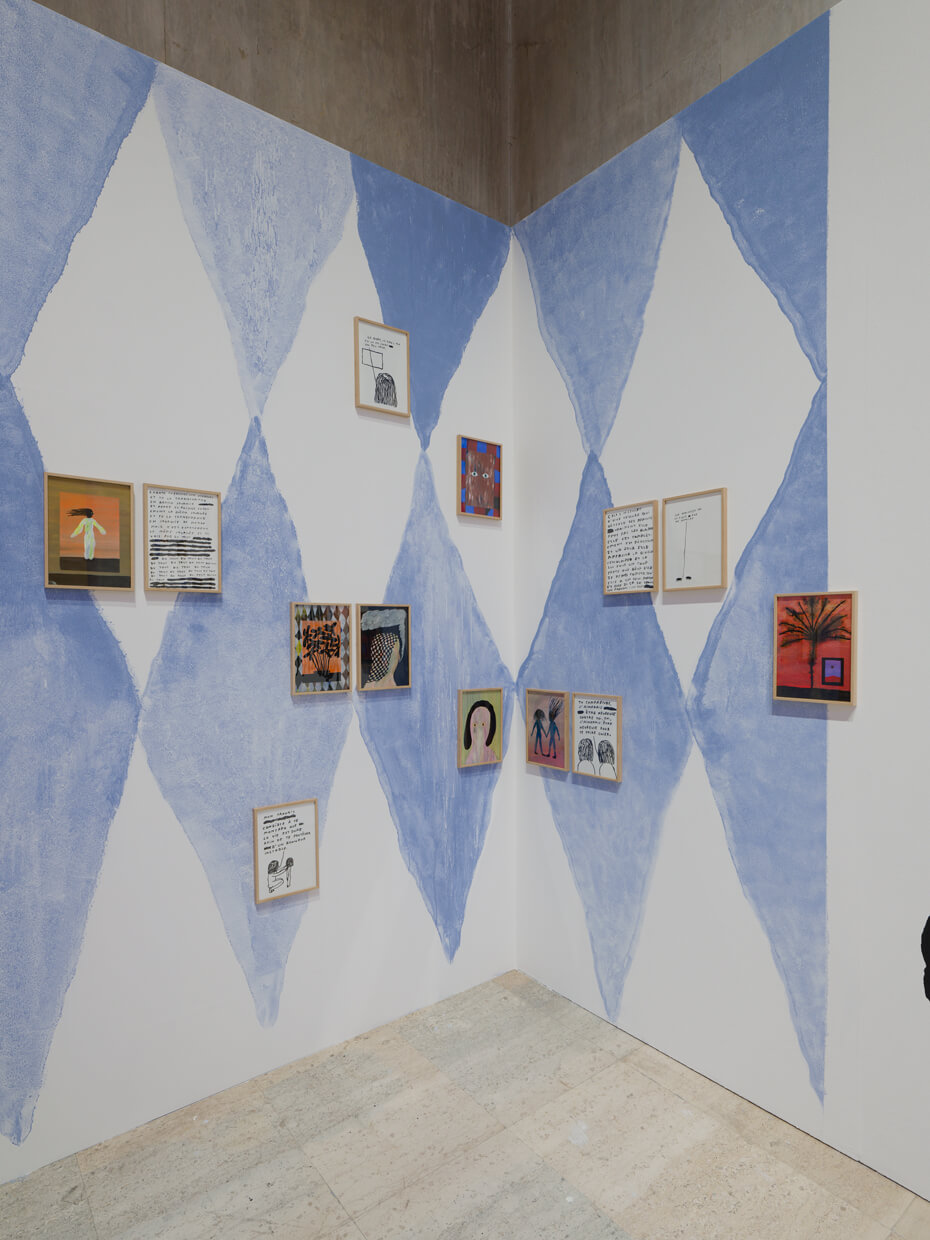 Un coin de galerie aux murs bleus à motifs de losanges où sont exposées des œuvres d'art et des illustrations encadrées et disposées selon une grille dispersée.