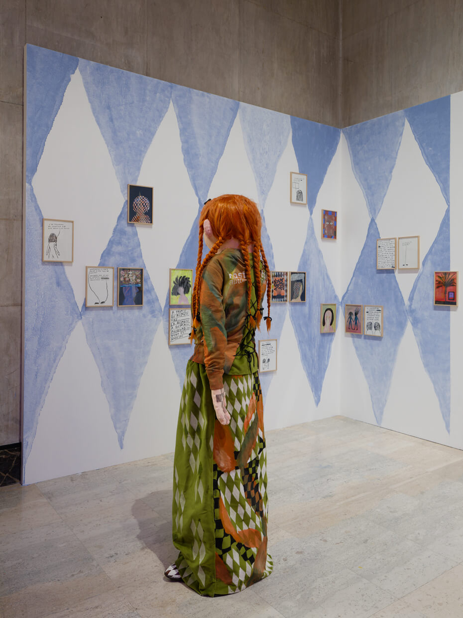 Un mannequin portant de longues tresses orange et une tenue verte à motifs se tient face à un mur orné de losanges bleus et de diverses œuvres d'art encadrées.