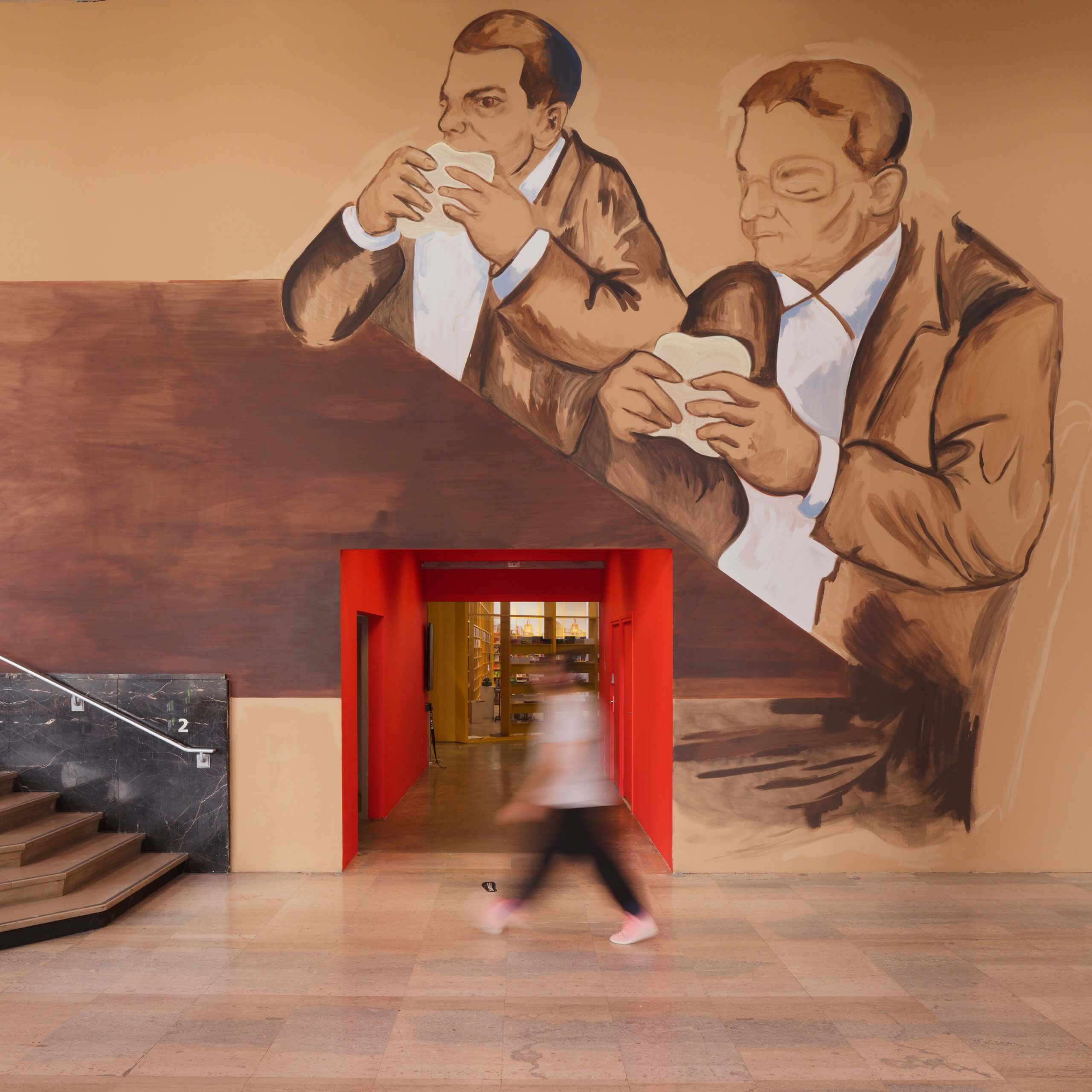 Grande fresque représentant deux hommes en costume en train de manger des sandwichs, peinte au-dessus et autour d'une porte ; une personne passe, floue en raison de son mouvement, à l'intérieur d'un bâtiment aux sols en marbre et doté d'un escalier.