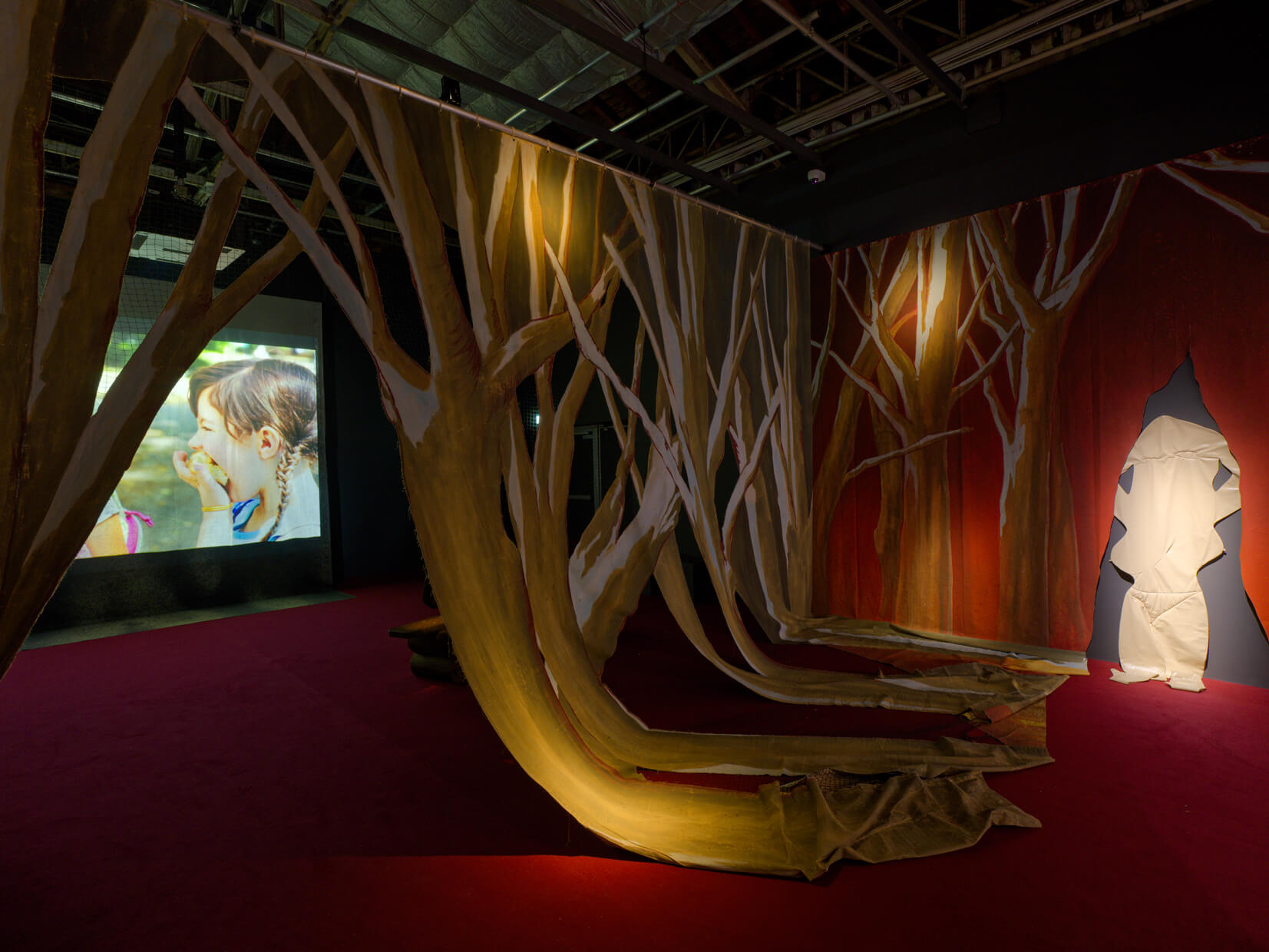 Room with large tree-like fabric structures, a video of a child and monkey on a screen, and a cutout doorway shaped like an irregular tree trunk; dim lighting, red floor.