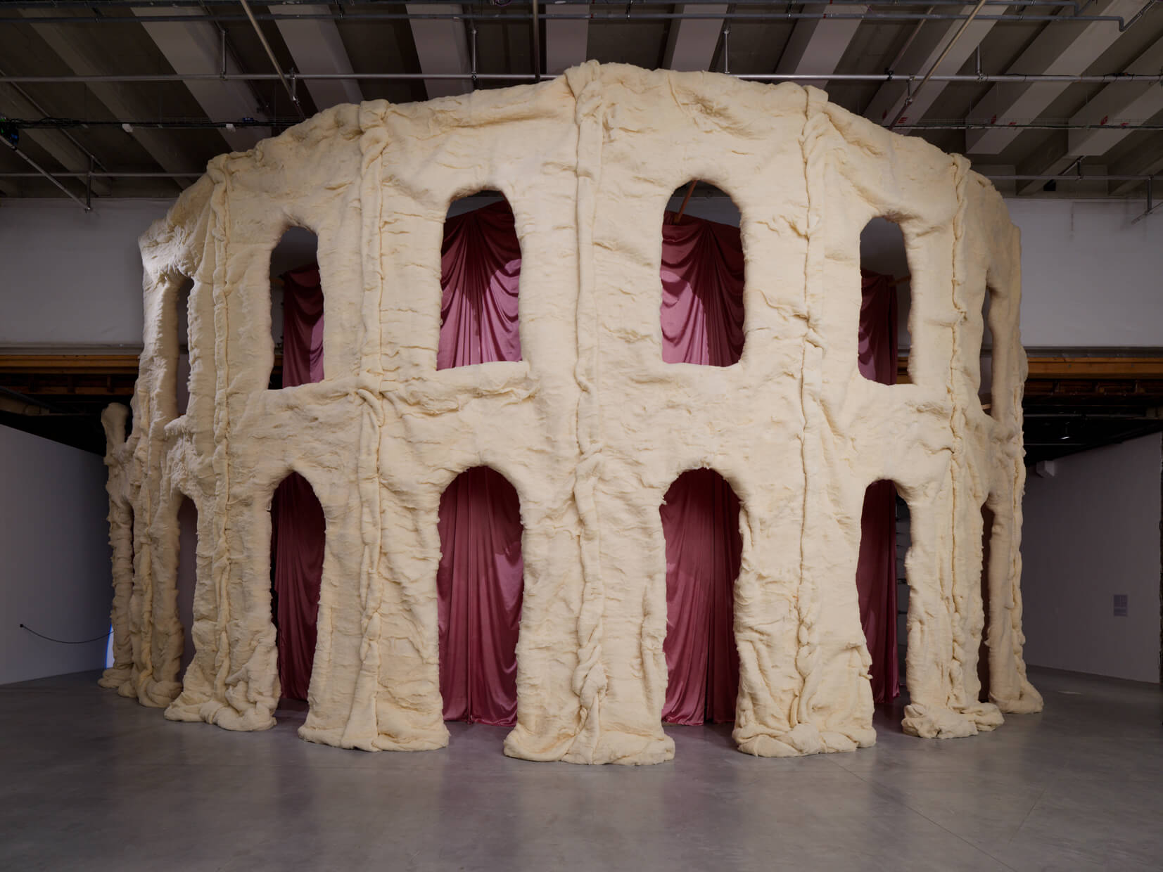 A large, circular sculpture resembling ancient ruins, with cream-colored, textured arches and pink fabric draped behind each opening, displayed in a gallery space.