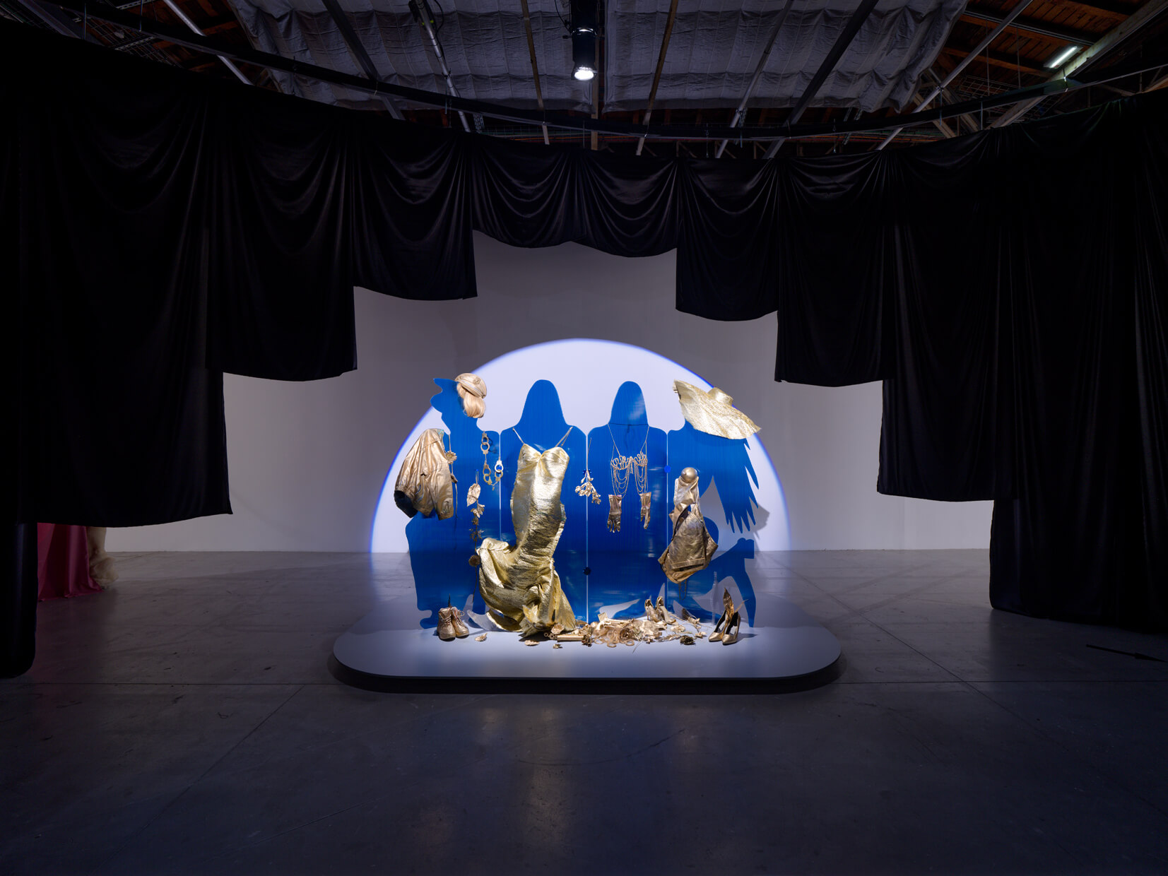 A blue silhouette backdrop displays gold sculptural objects and garments, illuminated by a spotlight, surrounded by black drapes in an industrial gallery space.