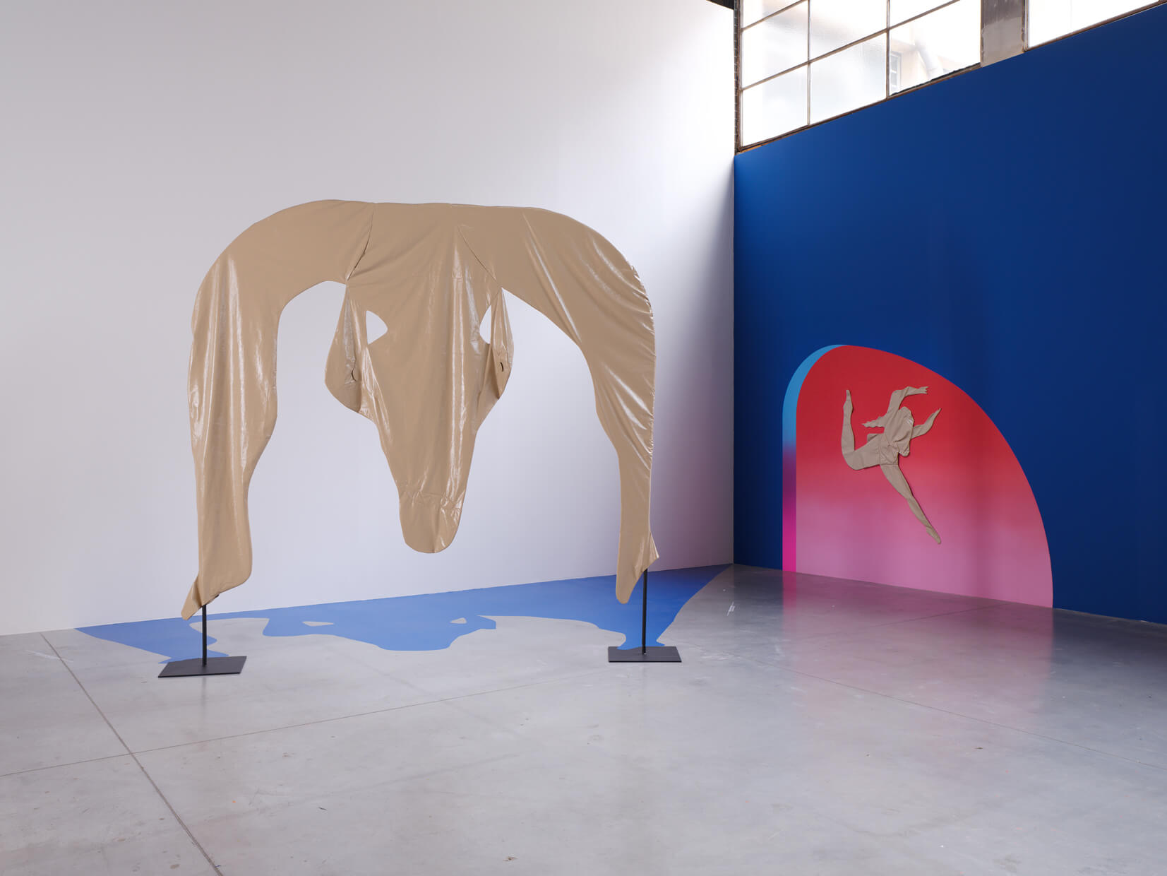 A tan fabric sculpture hangs on stands over a blue shadow shape; nearby, a wall mural shows a beige figure against a blue and pink gradient background.