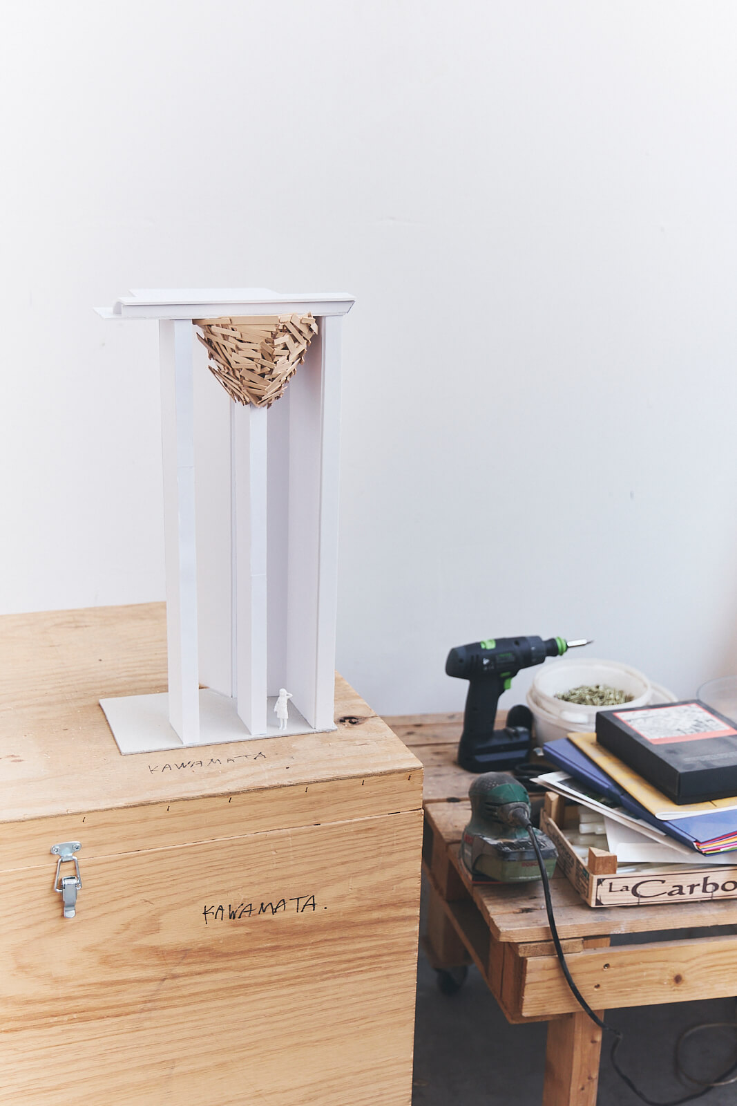 Une structure en bois avec un élément tissé est suspendue au sommet, placée sur une boîte à côté d'une table avec une perceuse, des livres et des outils.