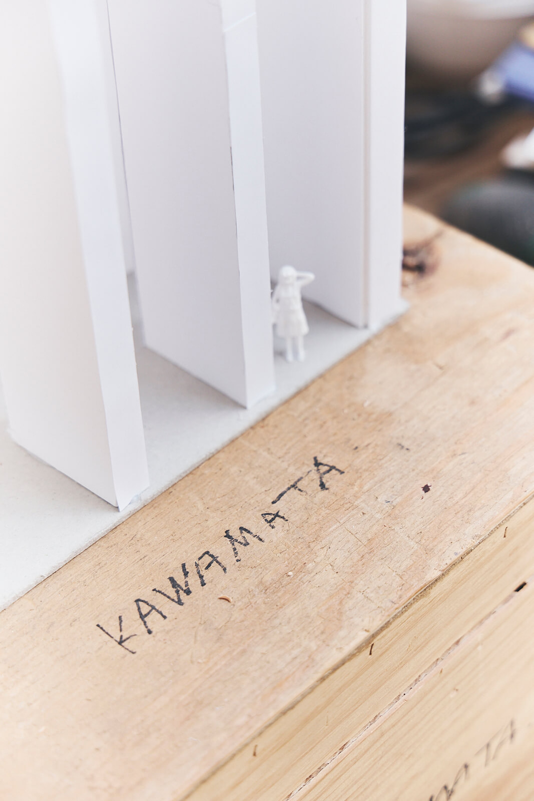 Une petite maquette architecturale avec des structures verticales blanches et une silhouette humaine miniature repose sur un socle en bois étiqueté KAWAMATA.