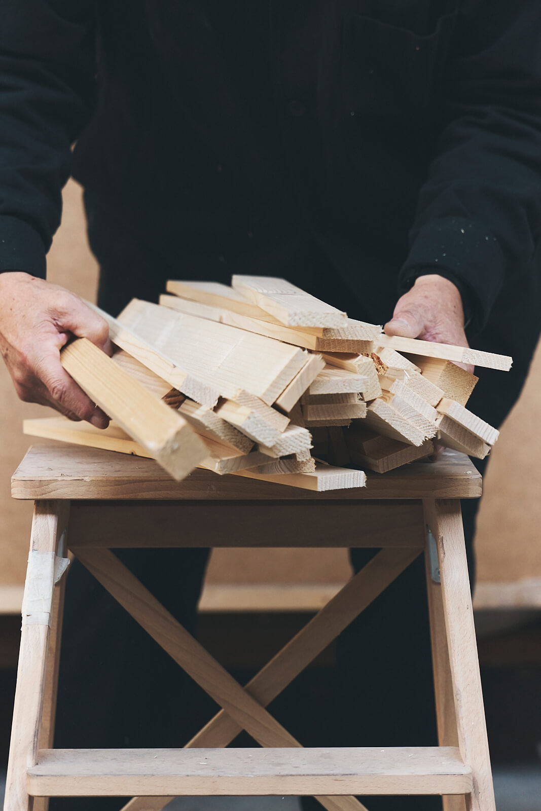 Une personne vêtue de vêtements sombres tient une pile de planches de bois au-dessus d'un petit tabouret en bois.
