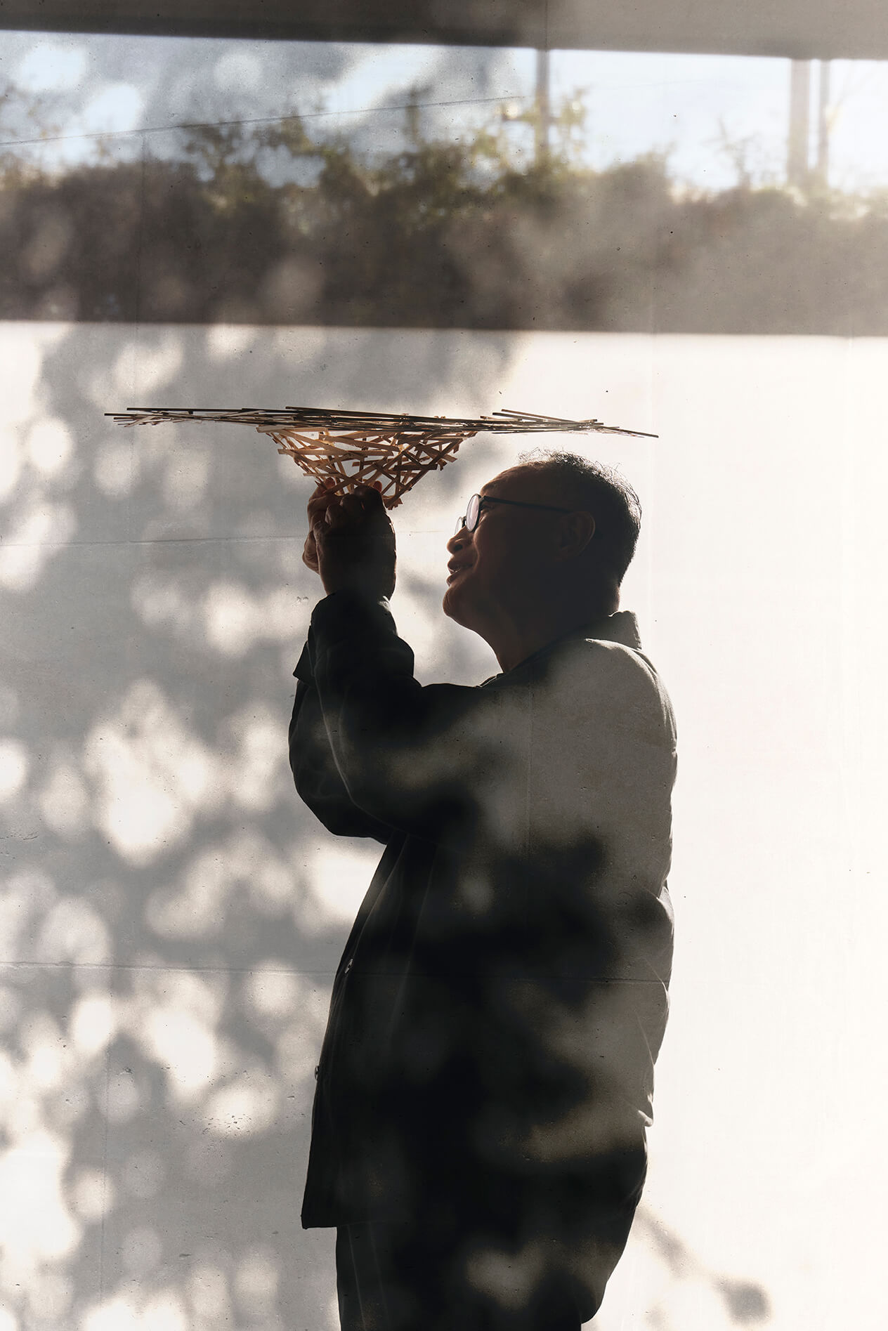Une personne de profil brandit une structure en bois en forme de treillis, l'examinant de près, debout, dans l'ombre et la lumière du soleil.
