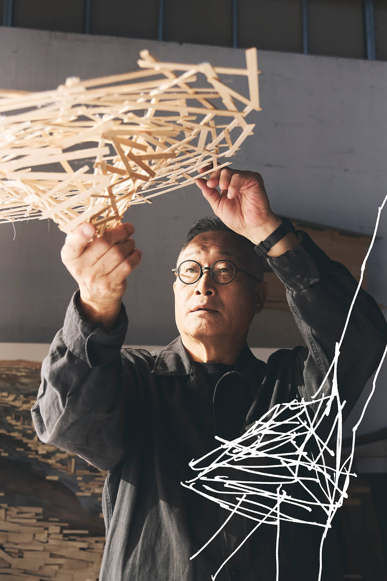 Un homme portant des lunettes et une veste sombre tient et examine une structure géométrique faite de fines baguettes de bois dans un espace intérieur bien éclairé.