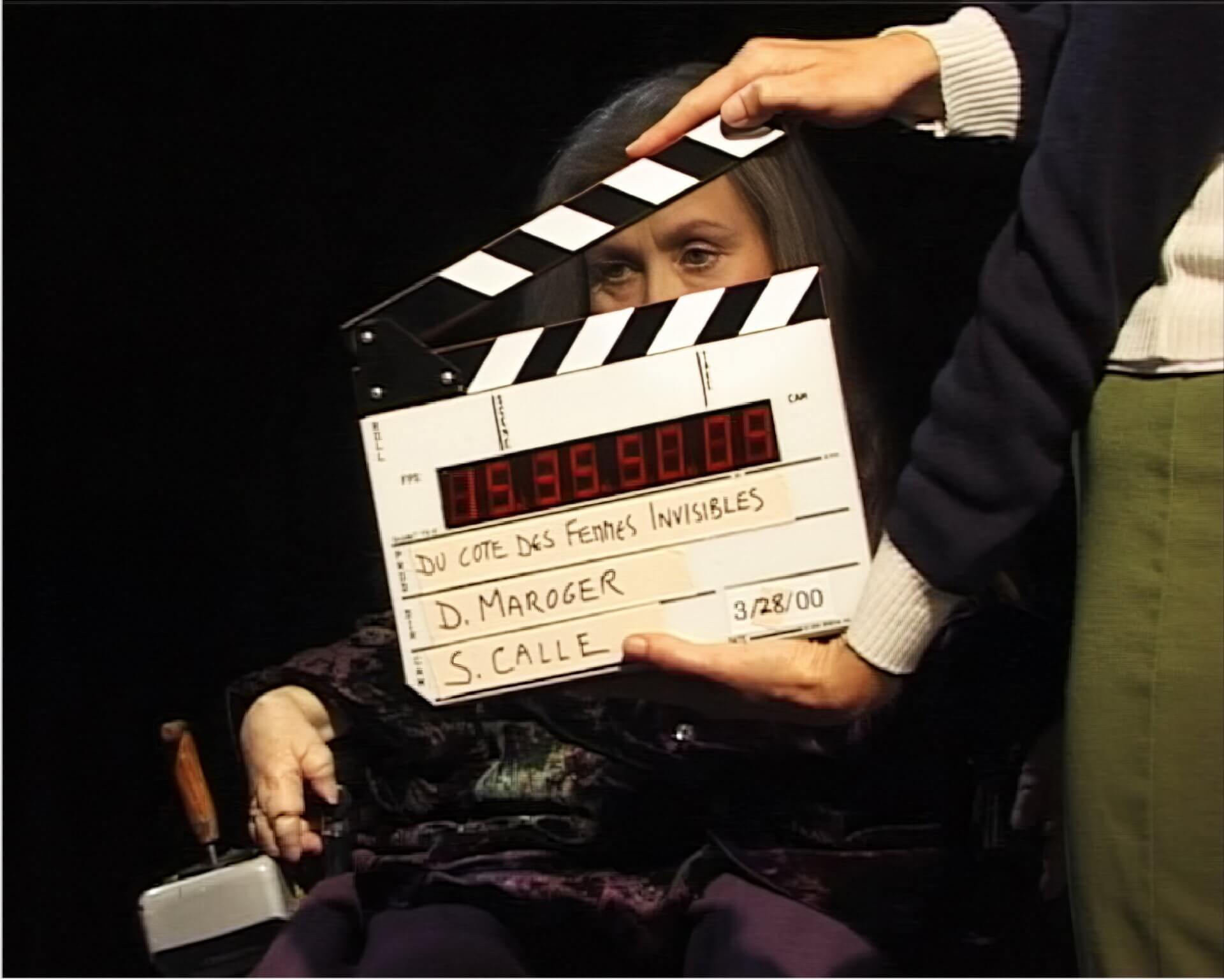 A person holds a film clapboard in front of an older woman sitting in a chair, partially obscuring her face; the slate reads D. Maroger, S. Calle, 3/28/00.