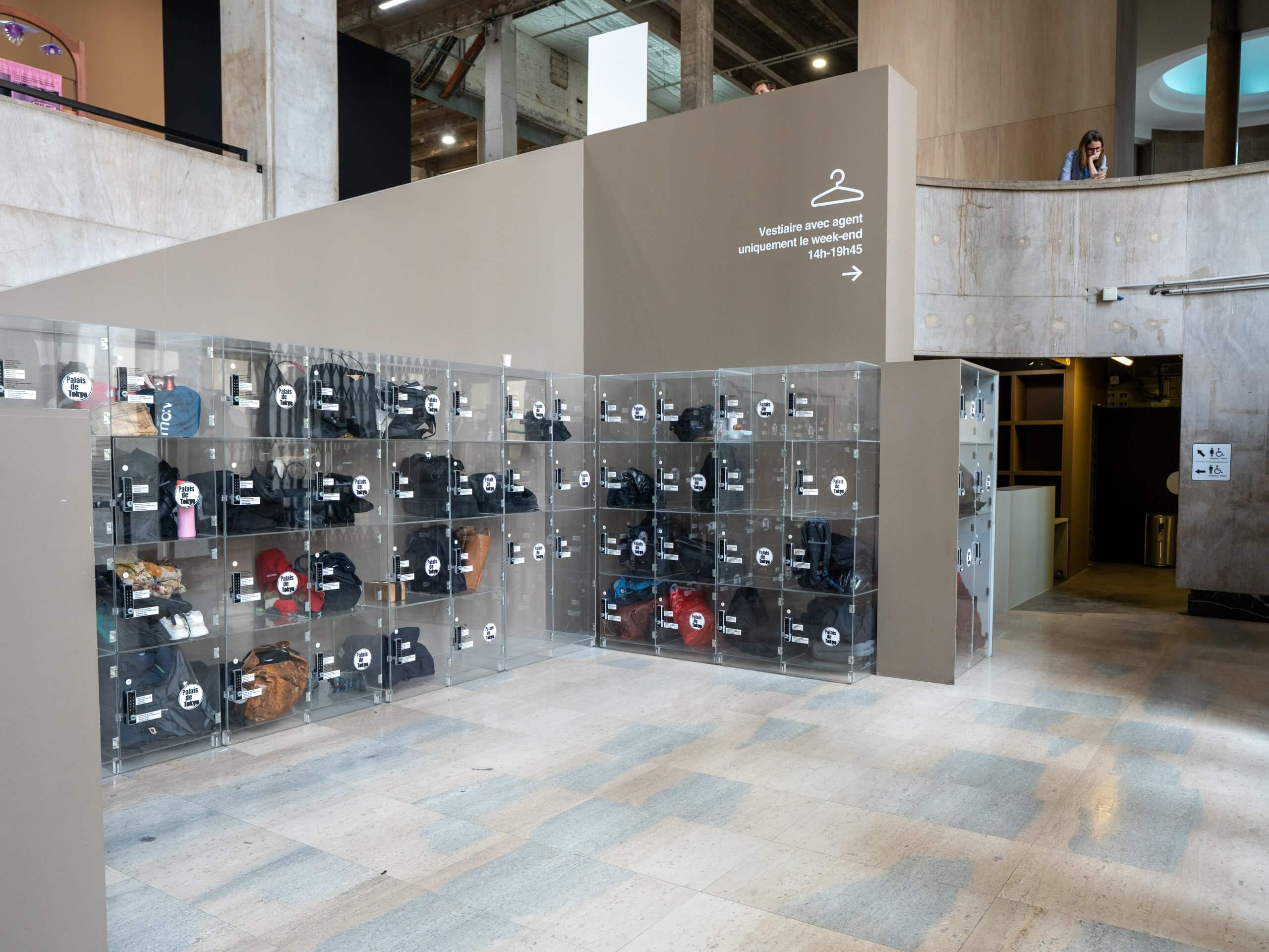 Un mur de casiers transparents remplis de sacs et d'objets personnels se trouve dans le hall d'un bâtiment moderne, avec un panneau indiquant un vestiaire à droite.
