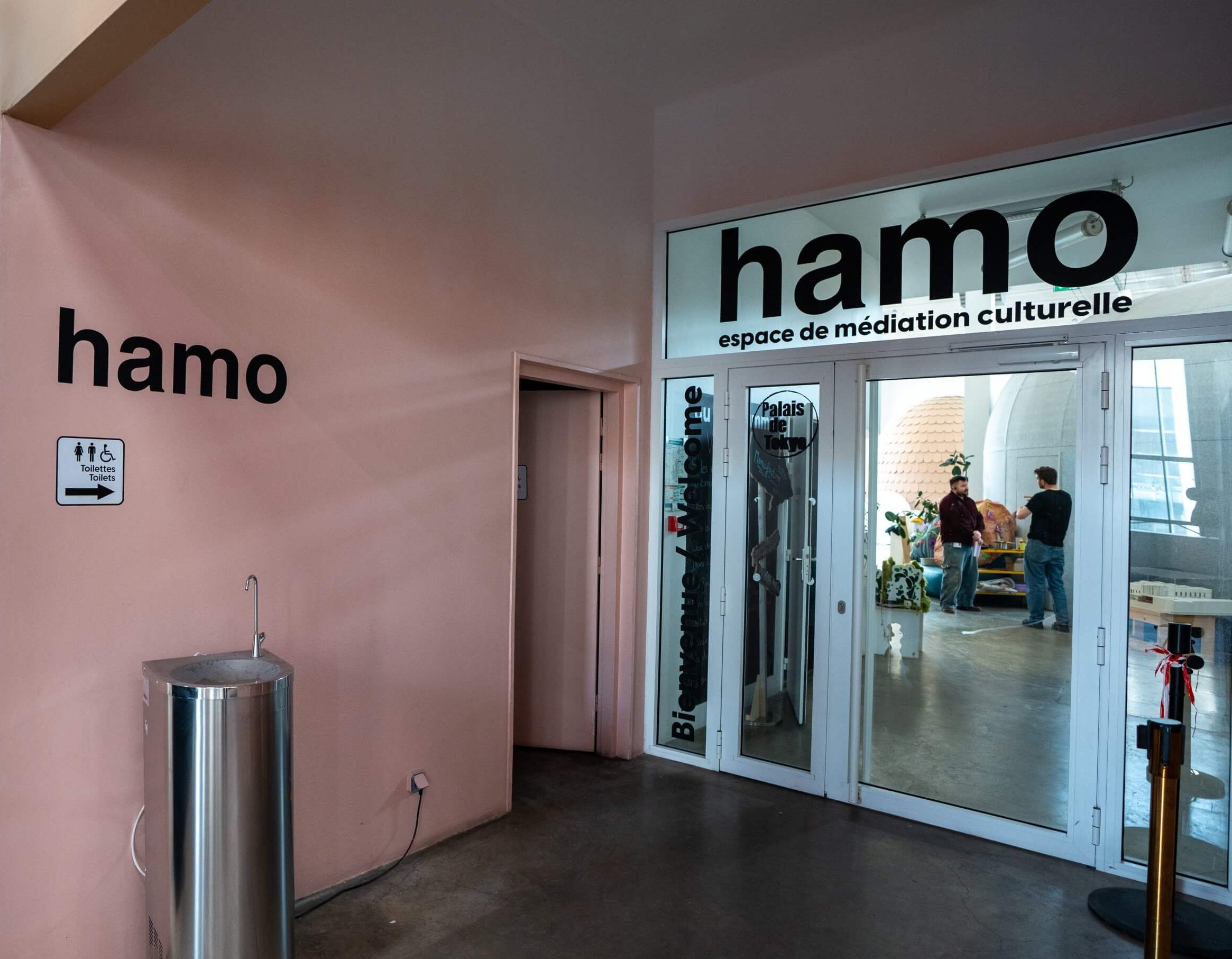 Entrée de l'espace de médiation culturelle hamo avec des portes vitrées, une signalétique, une fontaine et un panneau indiquant les toilettes ; plusieurs personnes se trouvent à l'intérieur de la salle.