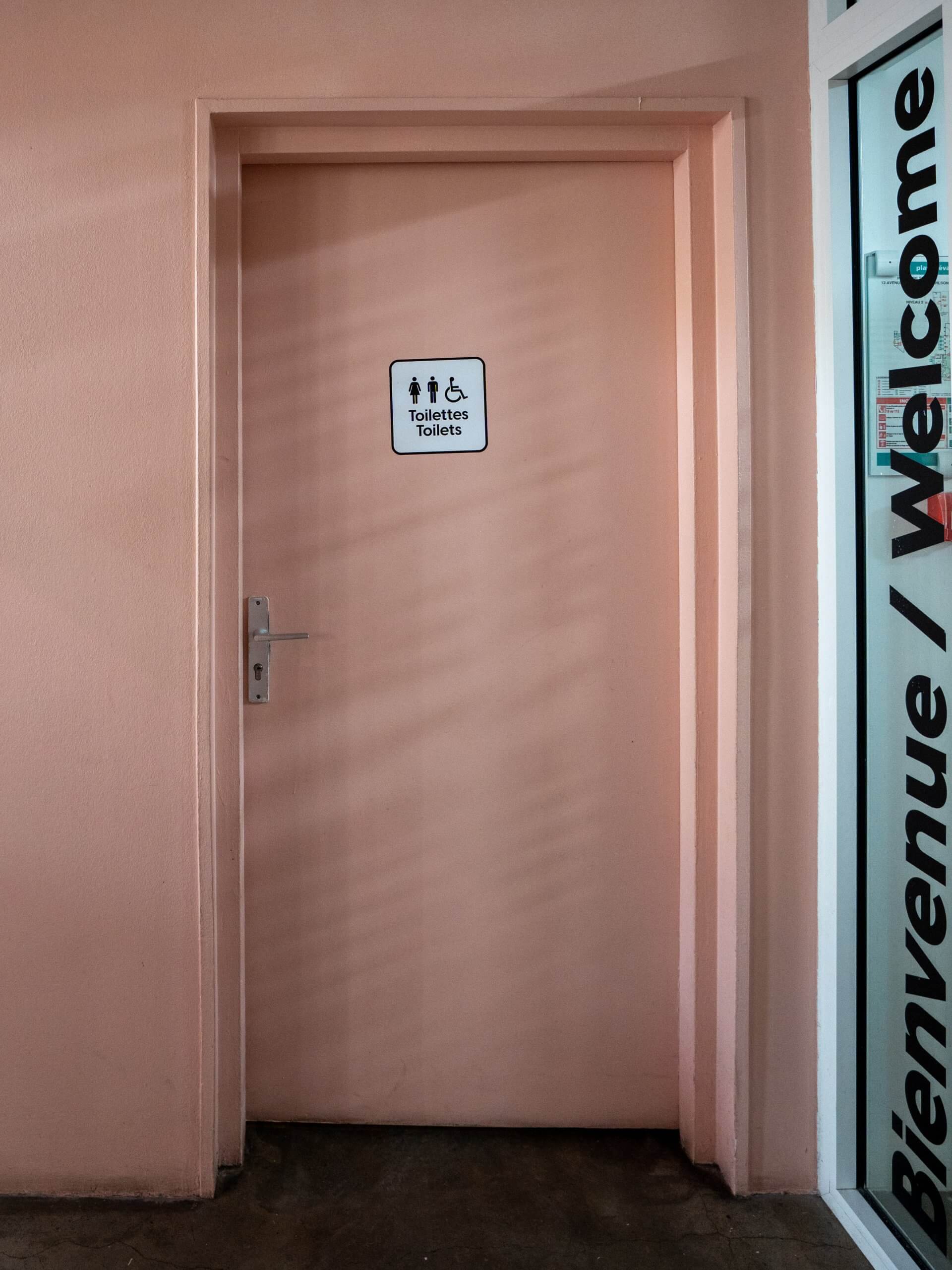Une porte de toilettes rose fermée avec un panneau indiquant des toilettes pour hommes, femmes et utilisateurs accessibles ; le texte Bienvenue / Welcome est partiellement visible sur le mur à côté.