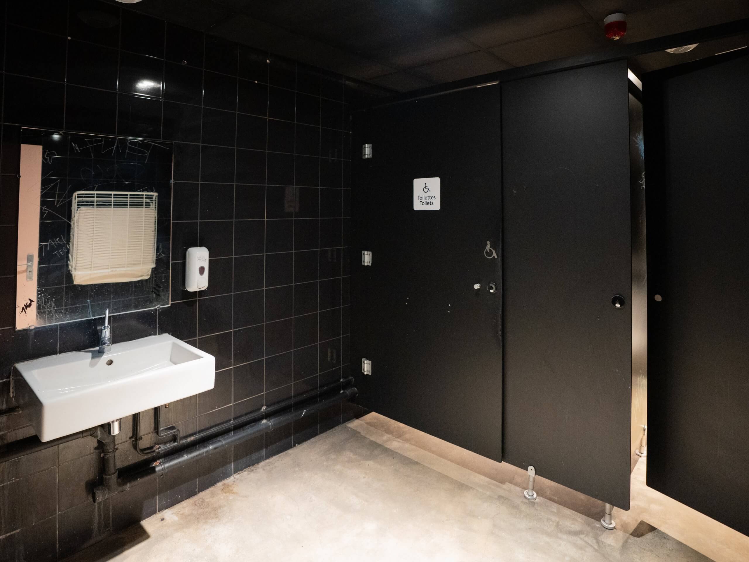 Toilettes publiques aux murs carrelés de noir, avec un lavabo blanc, un miroir, un distributeur de savon liquide et deux cabines de toilettes noires, dont l'une est indiquée comme toilettes accessibles.