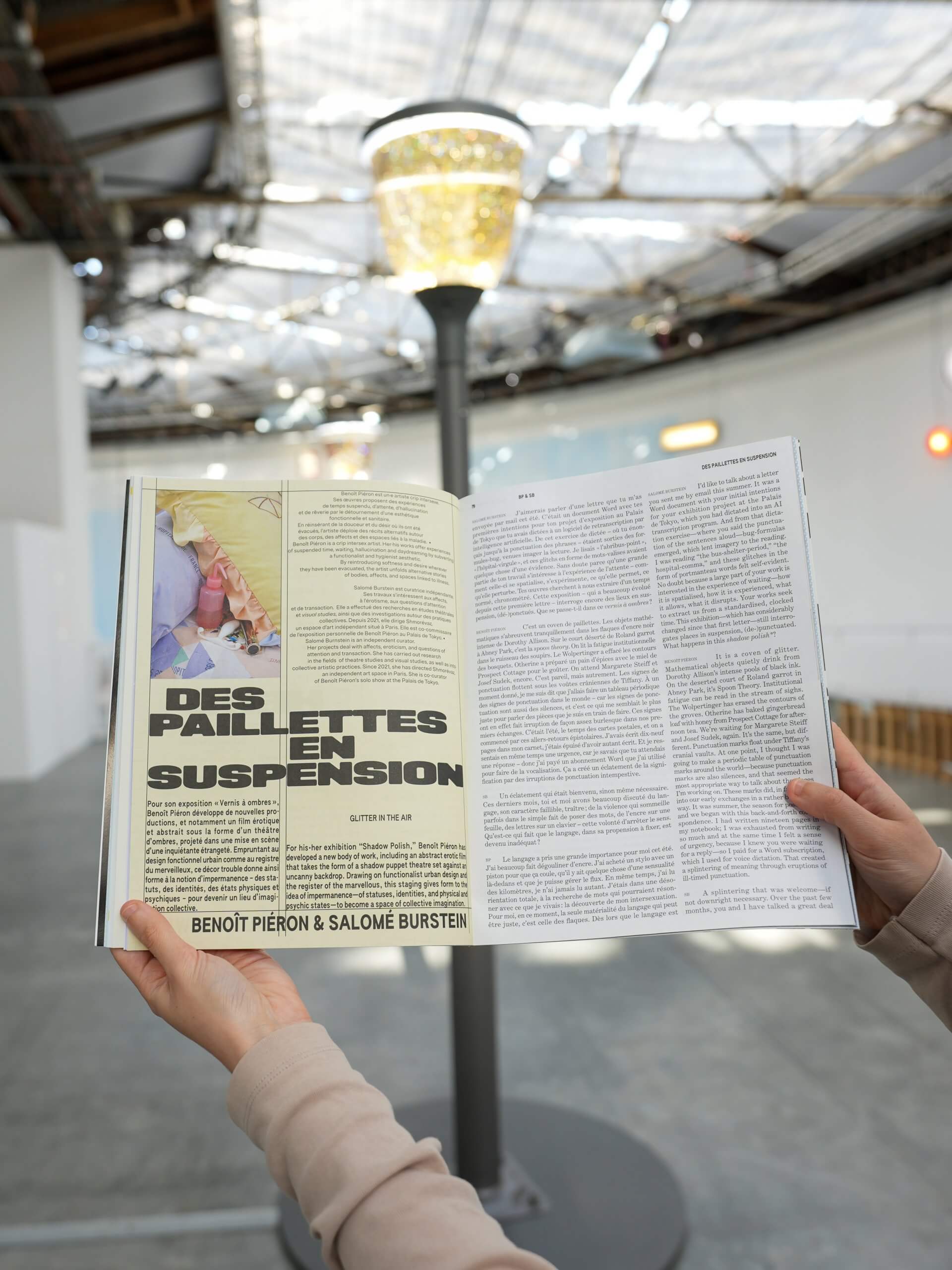 A person holds open a magazine or catalog displaying an article titled DES PAILLETTES EN SUSPENSION in a spacious, well-lit indoor space with a lamp post in the background.
