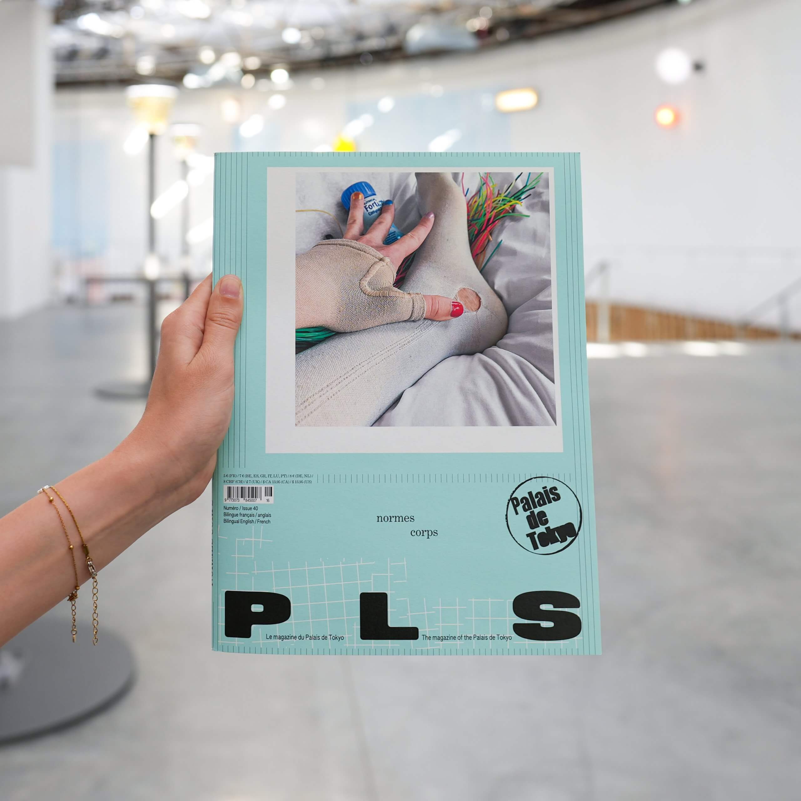 Une main tient un magazine vert pastel intitulé PLS, avec en couverture la photo d'une main portant une brassière beige et tenant des fleurs. L'arrière-plan est un espace intérieur moderne et aéré.