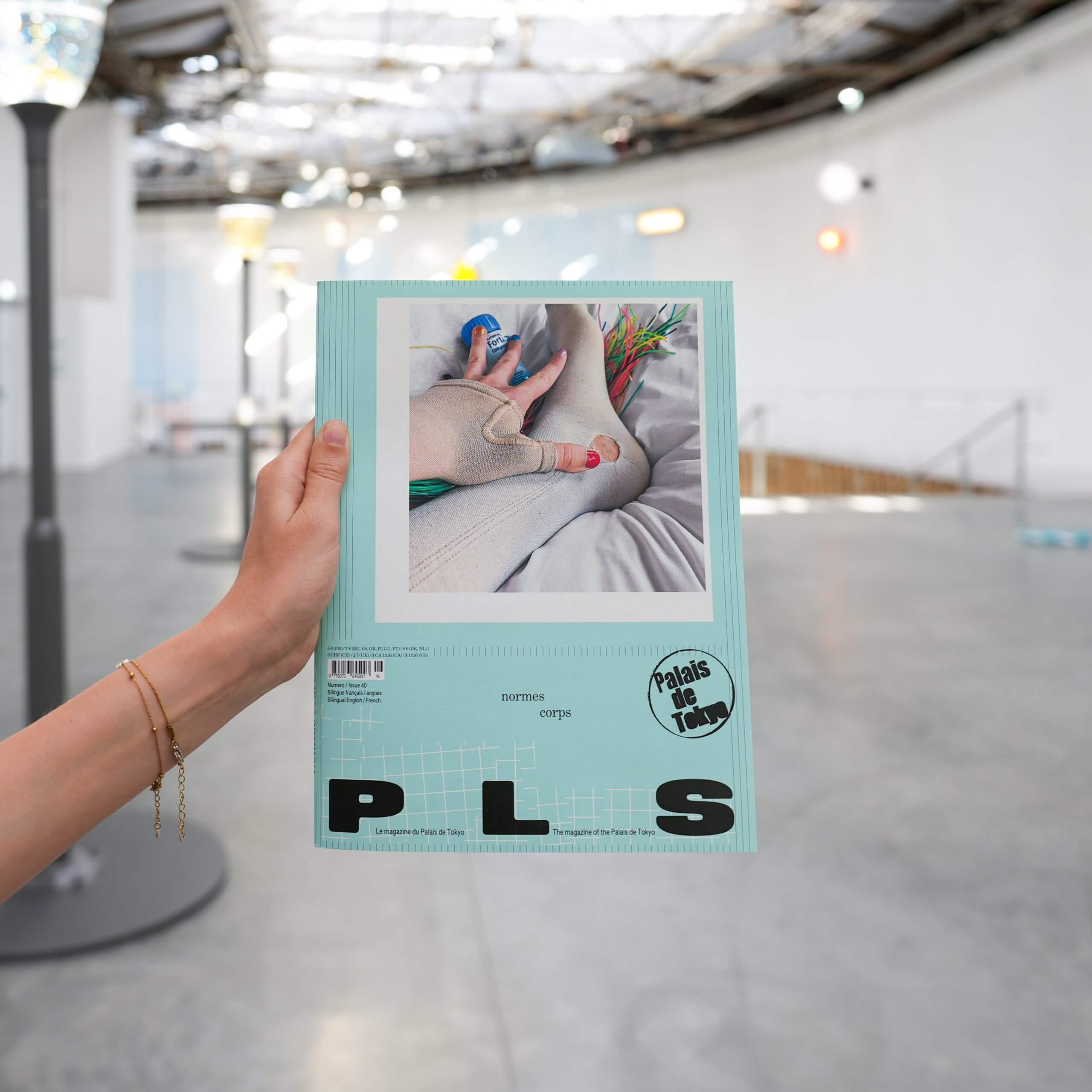 Une main tient un magazine intitulé PLS dont la couverture représente la main d'une personne tenant une fleur, à l'intérieur d'une galerie moderne et spacieuse.