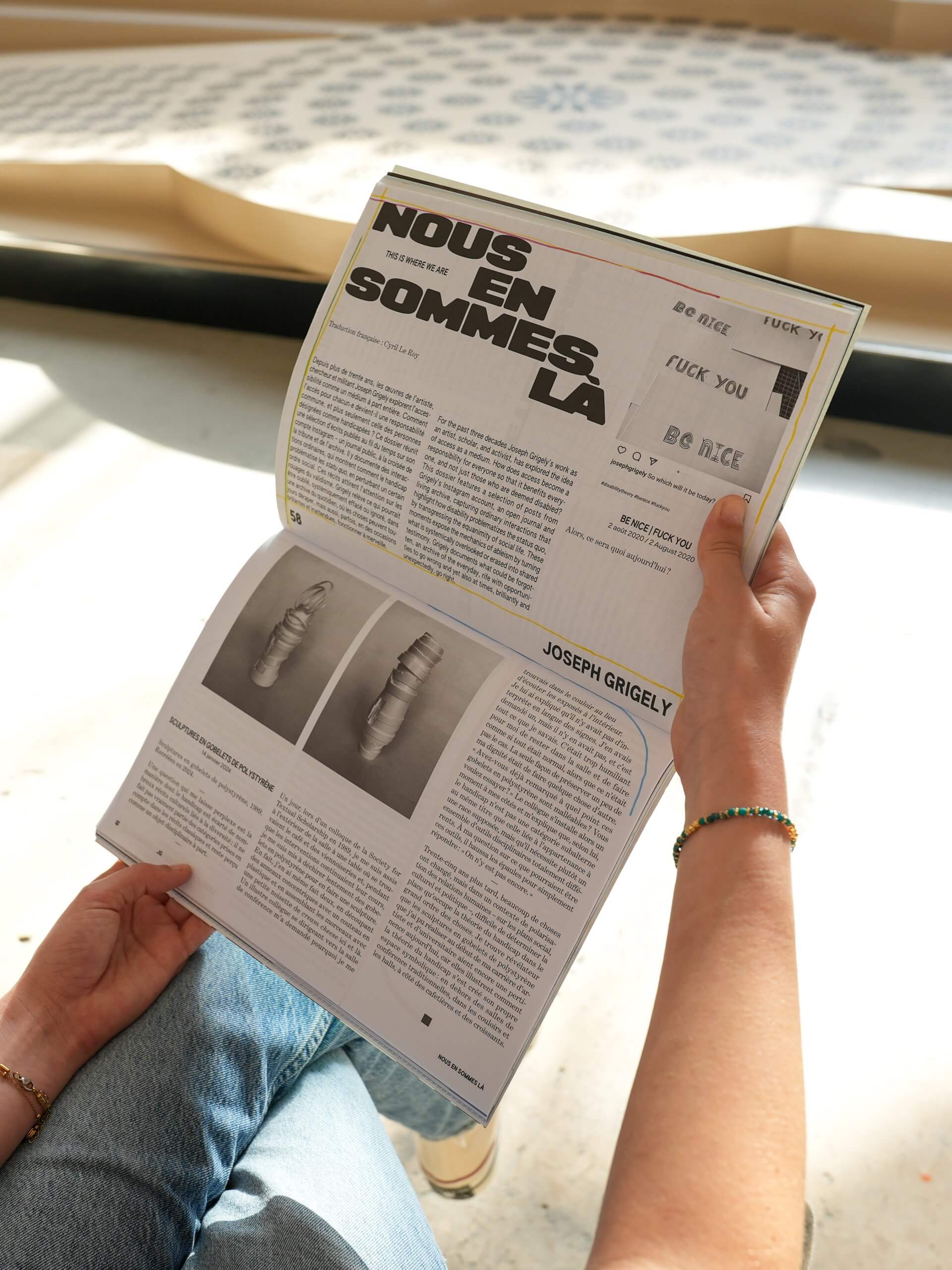 Person holding an open magazine featuring an article with bold French headline, NOUS EN SOMMES LA, and a section about artist Joseph Grigely with text and images of wrapped objects.