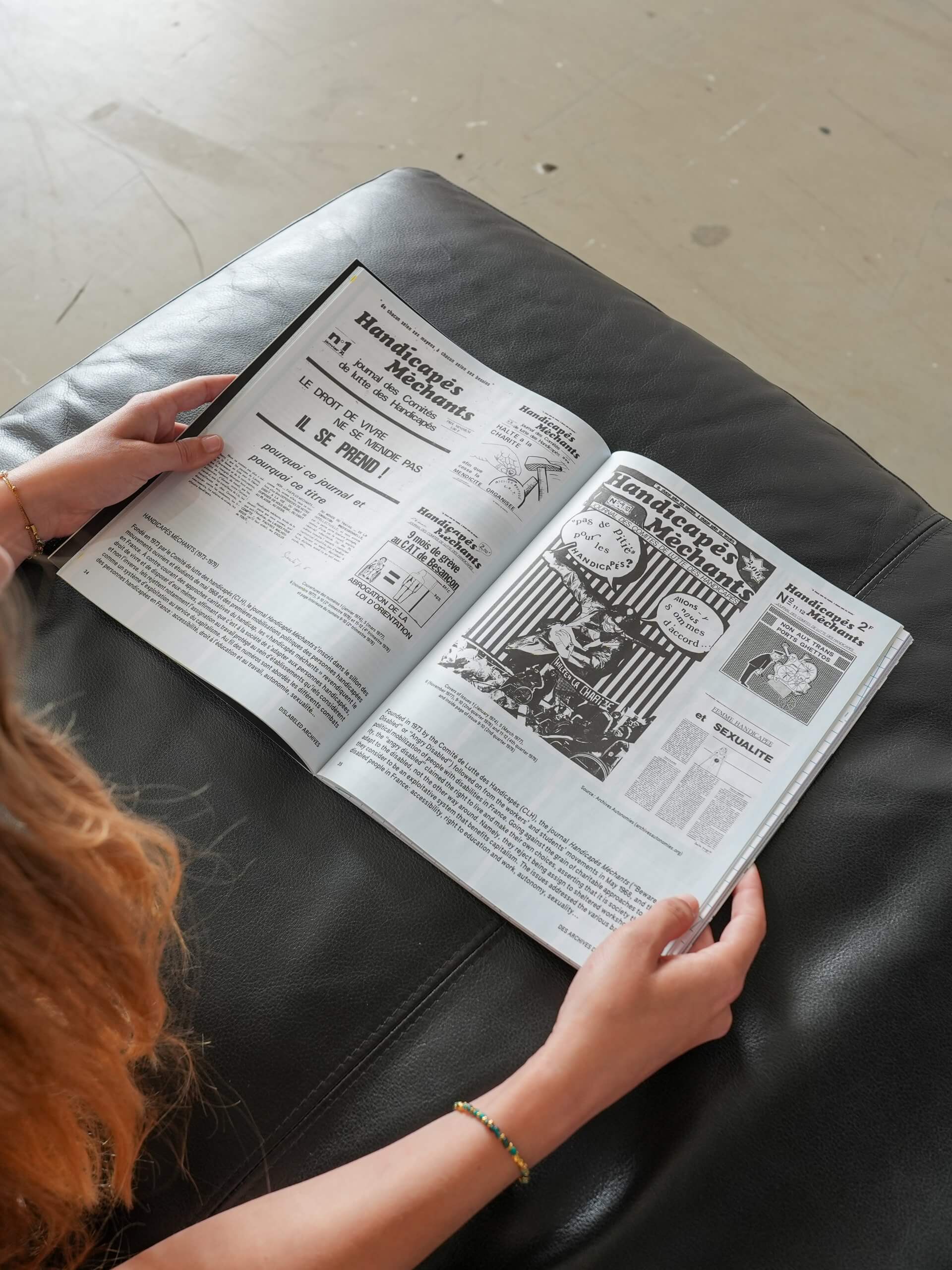 A person is sitting on a black leather sofa reading a magazine or newspaper with illustrated and text-heavy pages.