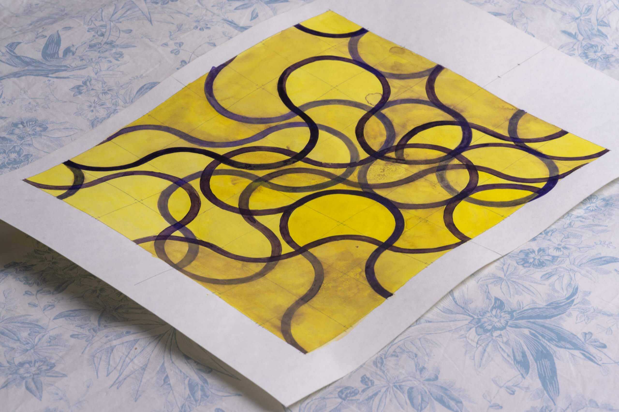 Une feuille de papier avec un motif abstrait dessiné à la main de lignes courbes foncées sur un fond jaune, placé sur une surface bleue à motifs floraux.