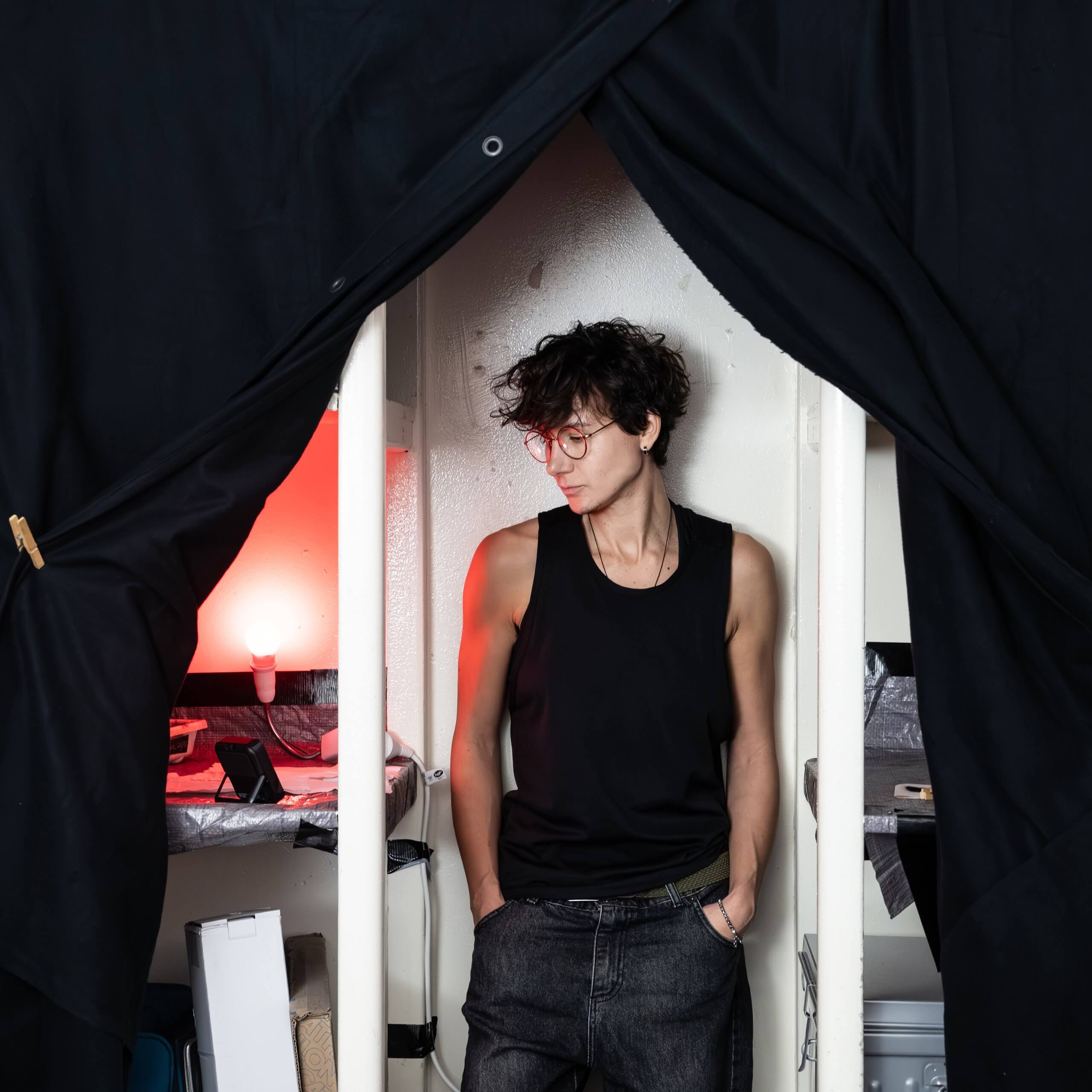 Une personne vêtue d'un débardeur noir et de lunettes se tient debout, les mains dans les poches, entre deux rideaux noirs, avec une lampe rouge et divers objets en arrière-plan.
