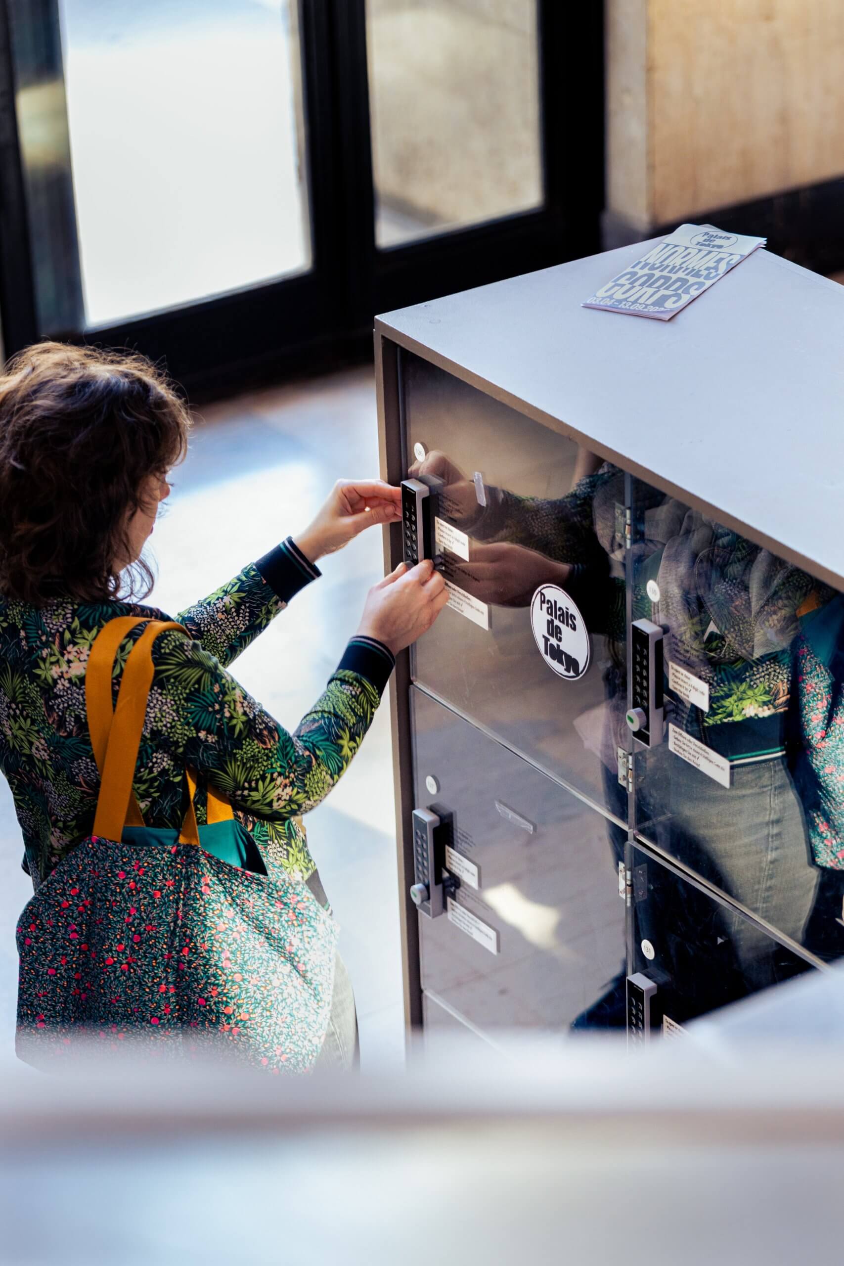 Une personne portant une veste à motifs et un sac fourre-tout utilise une clé pour ouvrir un casier dans un espace public intérieur.