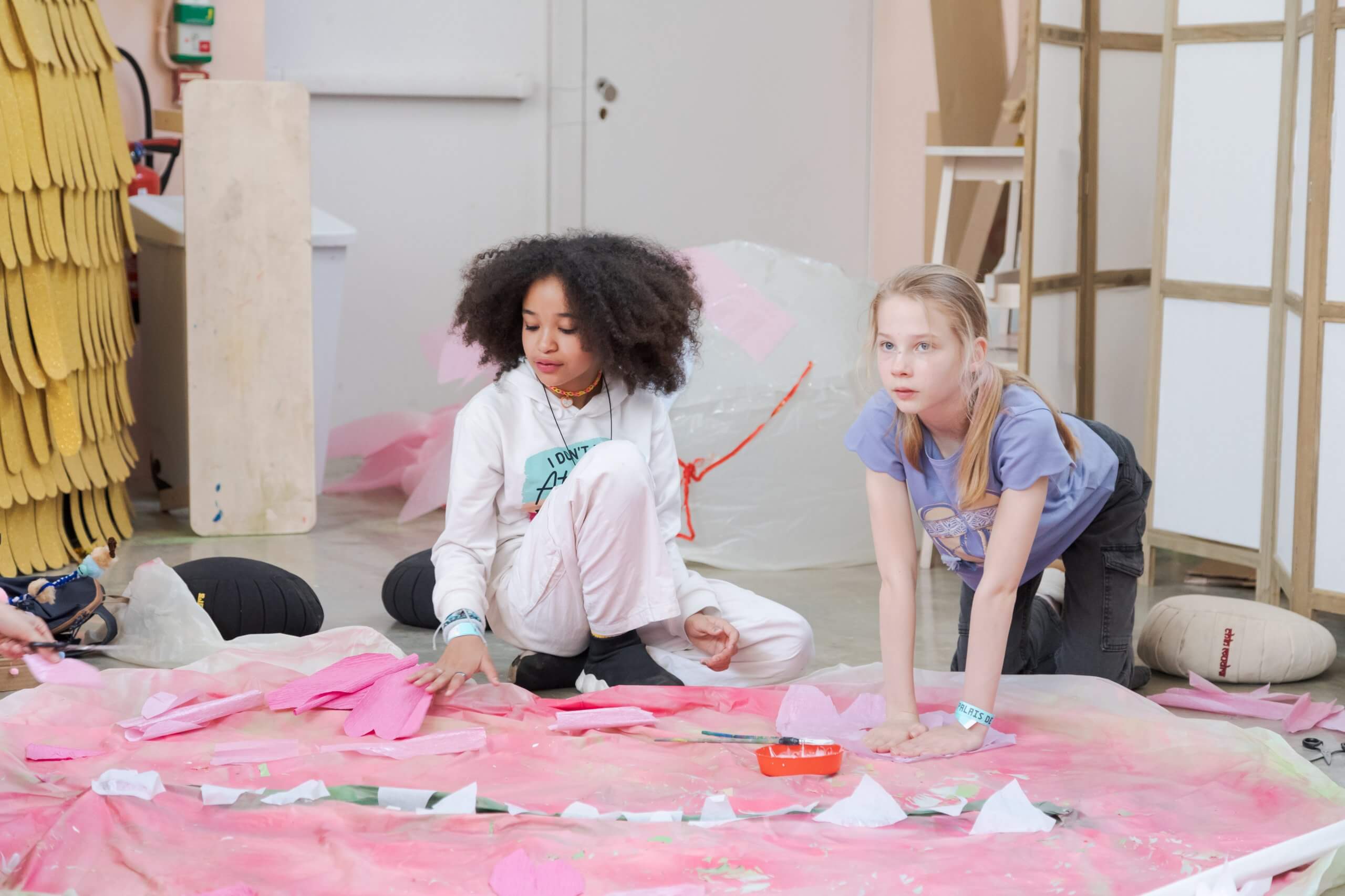 Deux filles sont agenouillées sur le sol, travaillant avec du papier rose et du matériel de bricolage étalé sur une grande feuille rose dans un espace intérieur.
