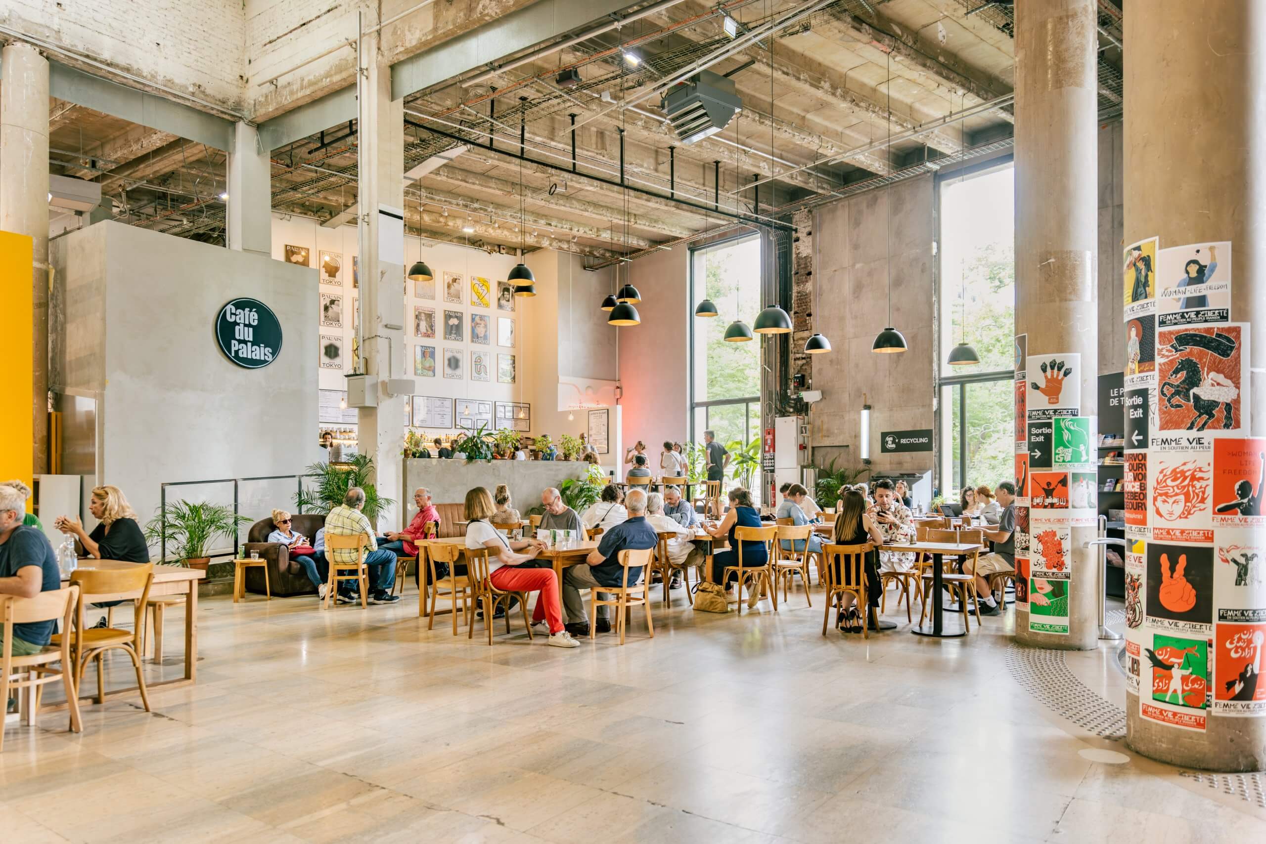 Café spacieux avec de hauts plafonds et de grandes fenêtres, rempli de gens assis à des tables et en train de socialiser ; des affiches colorées couvrent une colonne à proximité.