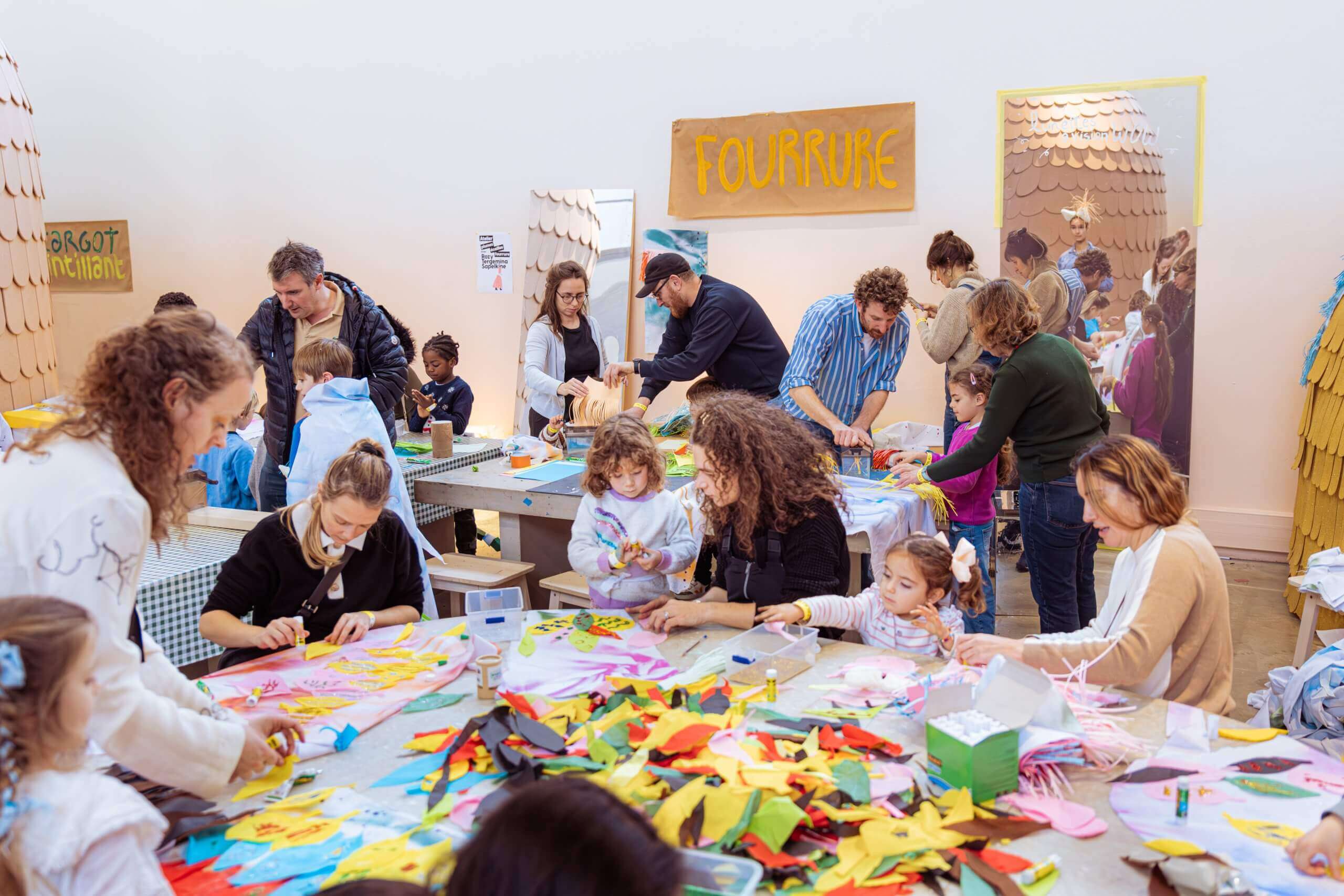 Des adultes et des enfants sont assis et debout autour de tables, engagés dans des activités artistiques et artisanales colorées dans une pièce lumineuse et animée.