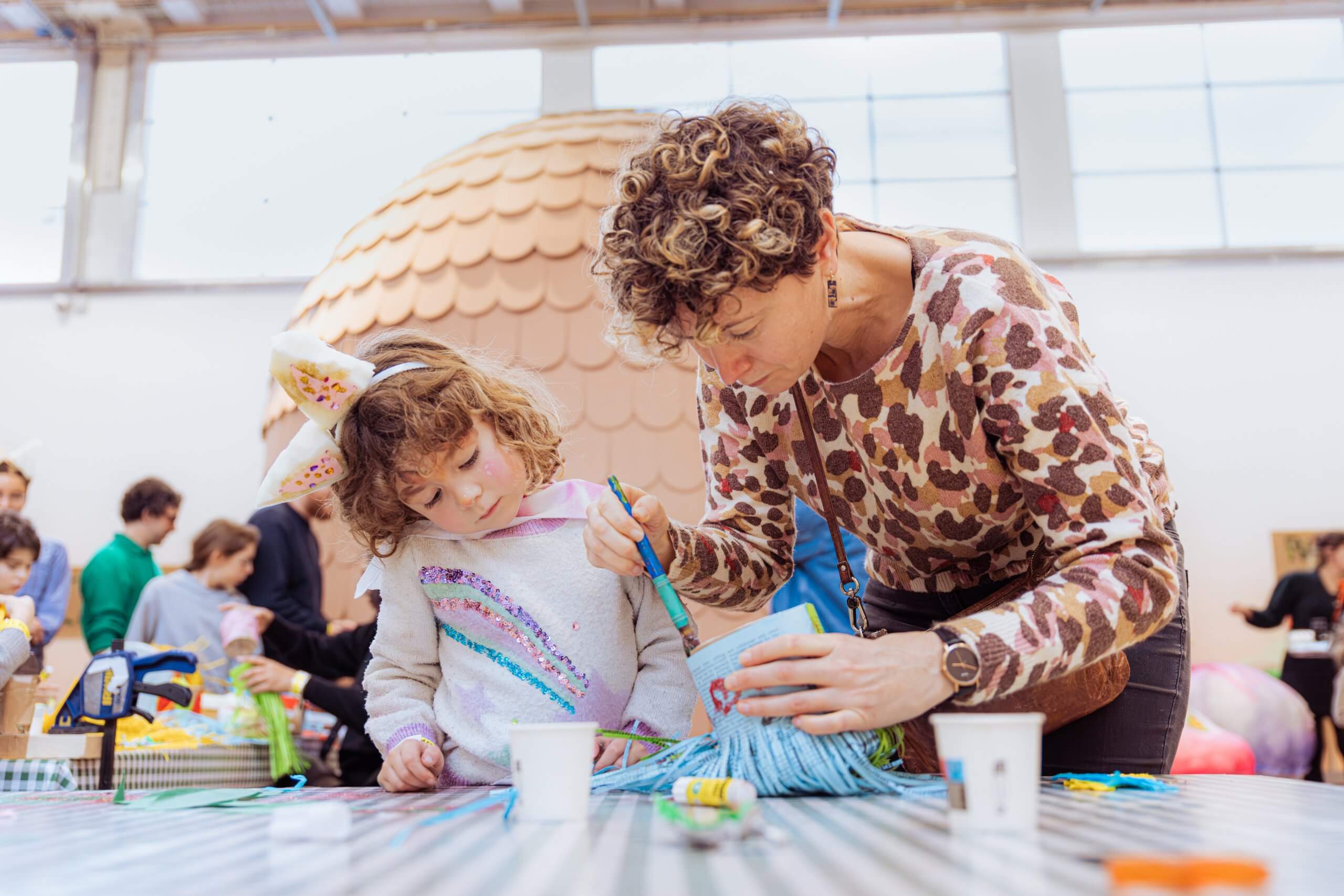 Un adulte aide un enfant à réaliser un projet d'artisanat à une table, en utilisant du fil et de la peinture. D'autres personnes sont visibles à l'arrière-plan dans un espace intérieur lumineux.