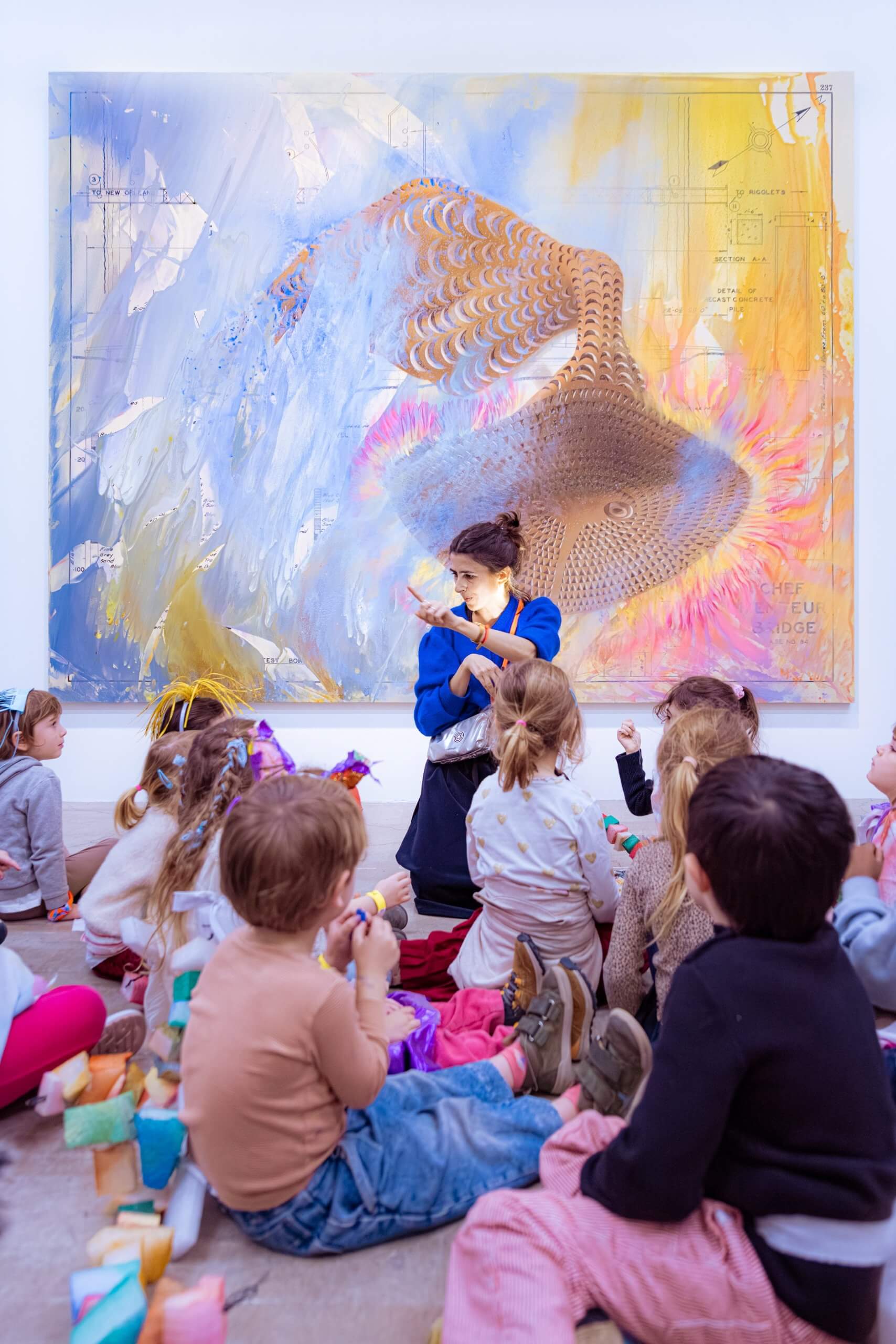 Dans une galerie, un groupe de jeunes enfants est assis par terre, face à une femme qui tient un objet, devant une grande peinture abstraite et colorée.