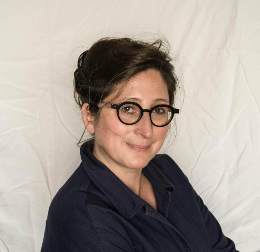 Une femme aux cheveux noirs coiffés en chignon, portant des lunettes noires rondes et une chemise marine, est assise devant un fond blanc texturé.