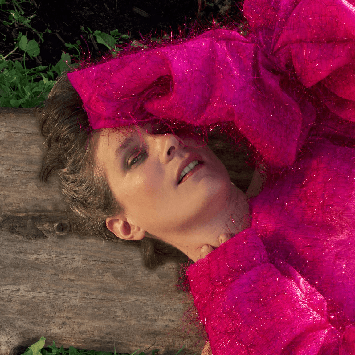 Personne vêtue d'une tenue rose vif et texturée, allongée sur des planches de bois, un bras couvrant une partie de son visage, entourée de plantes vertes.