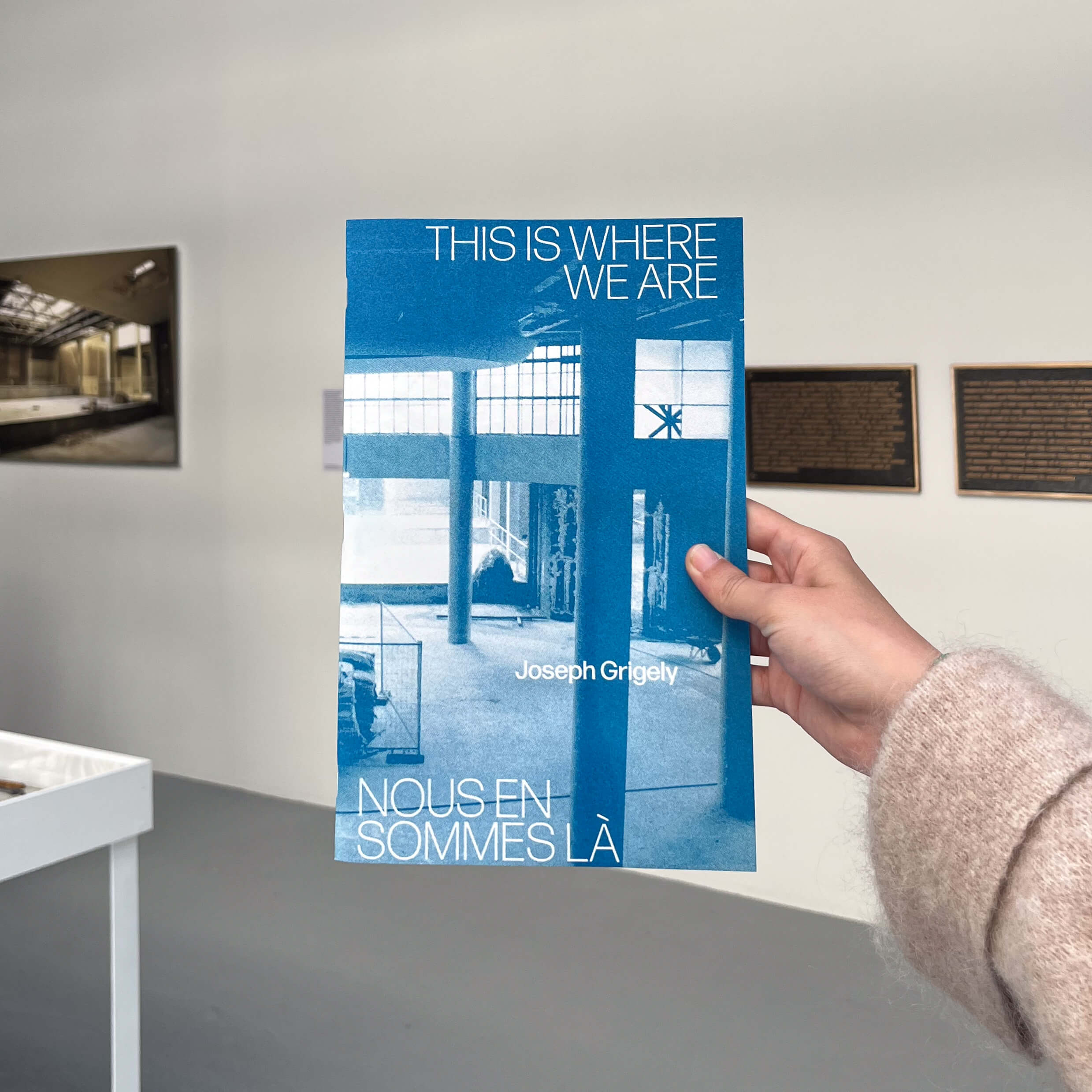 Une main tient un livret d'exposition bleu et blanc intitulé This Is Where We Are / Nous En Sommes Là de Joseph Grigely dans une galerie minimaliste.