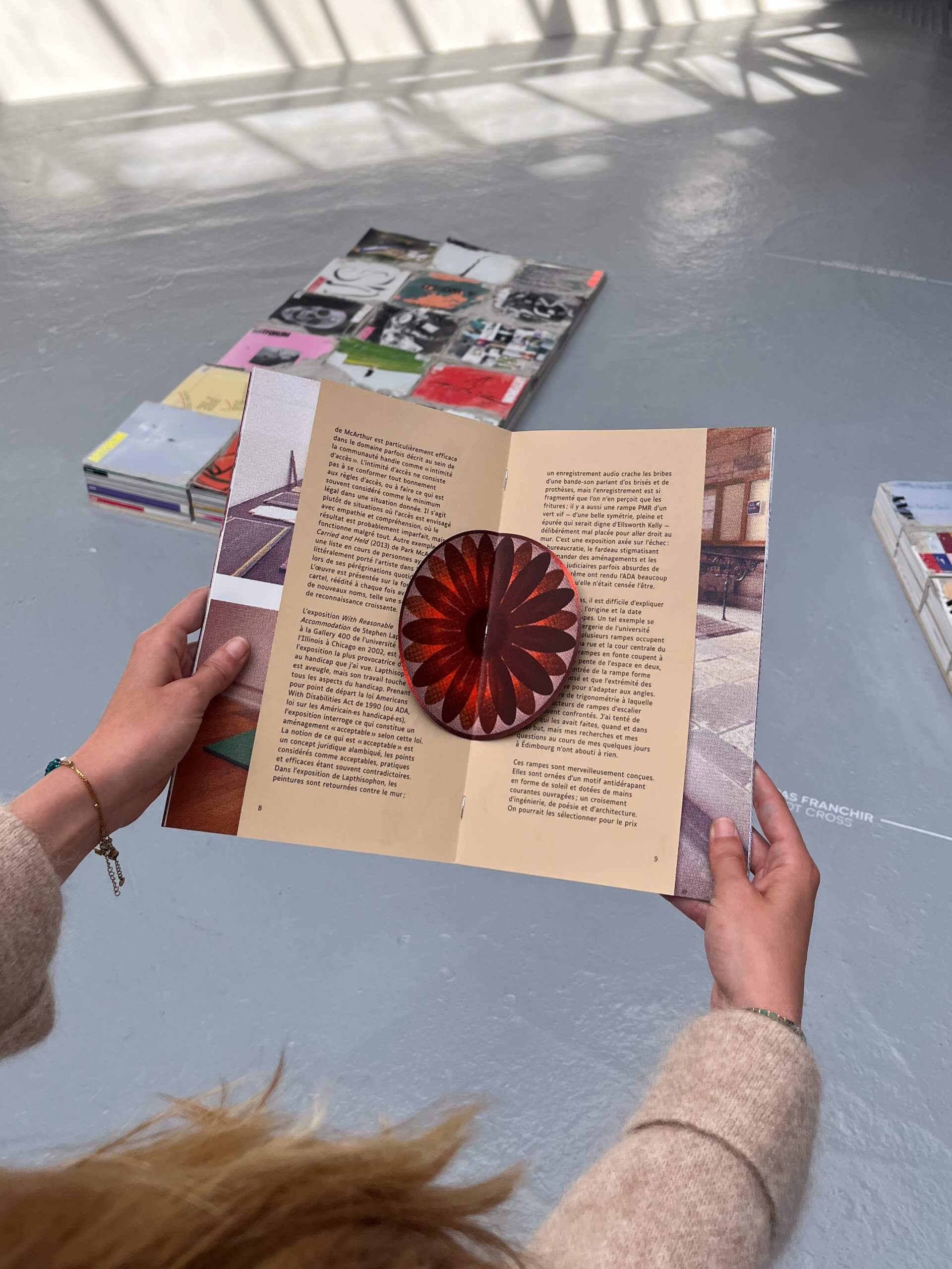 Une personne tient un livre ouvert avec, au centre, un dessin circulaire en papier 3D de couleur marron ; des piles de livres sont disposées sur un sol gris à l'arrière-plan.