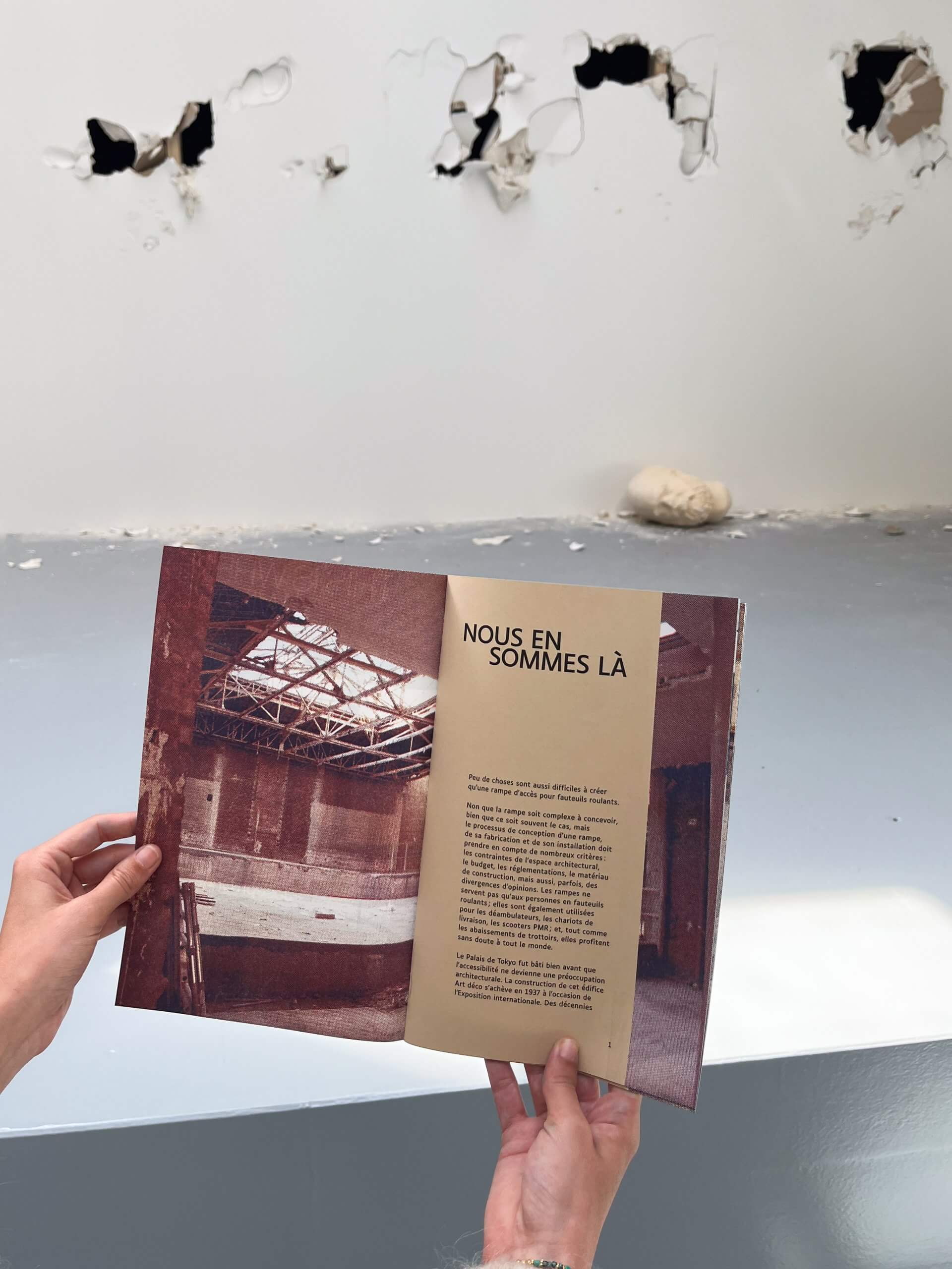 Une personne tient ouvert un livre sur lequel figure une photo d'un espace industriel vide et un texte en français, avec en arrière-plan un mur troué.