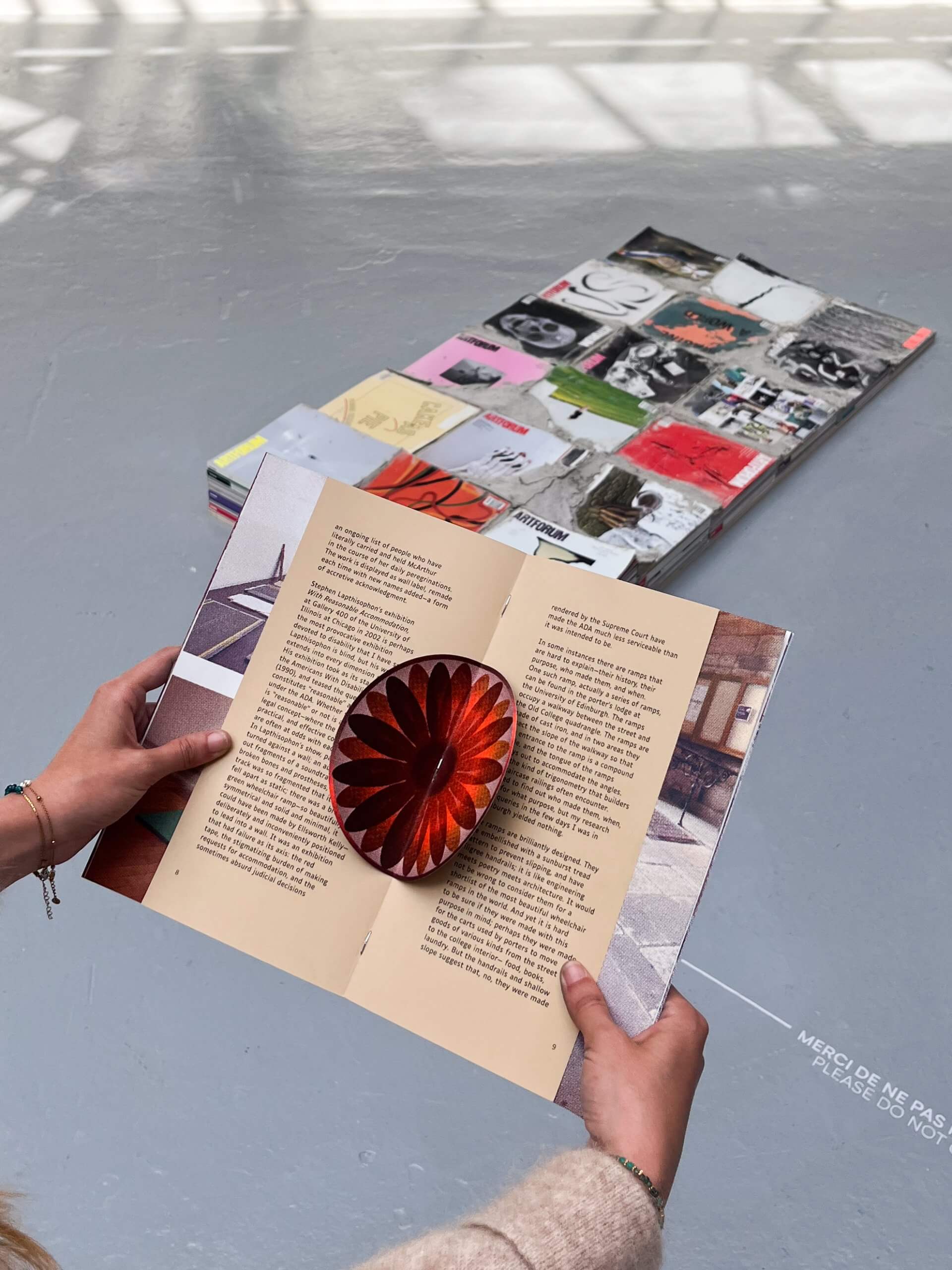 A person holds an open book with a red, circular 3D object in the center; several magazines and books are arranged on the floor in the background.