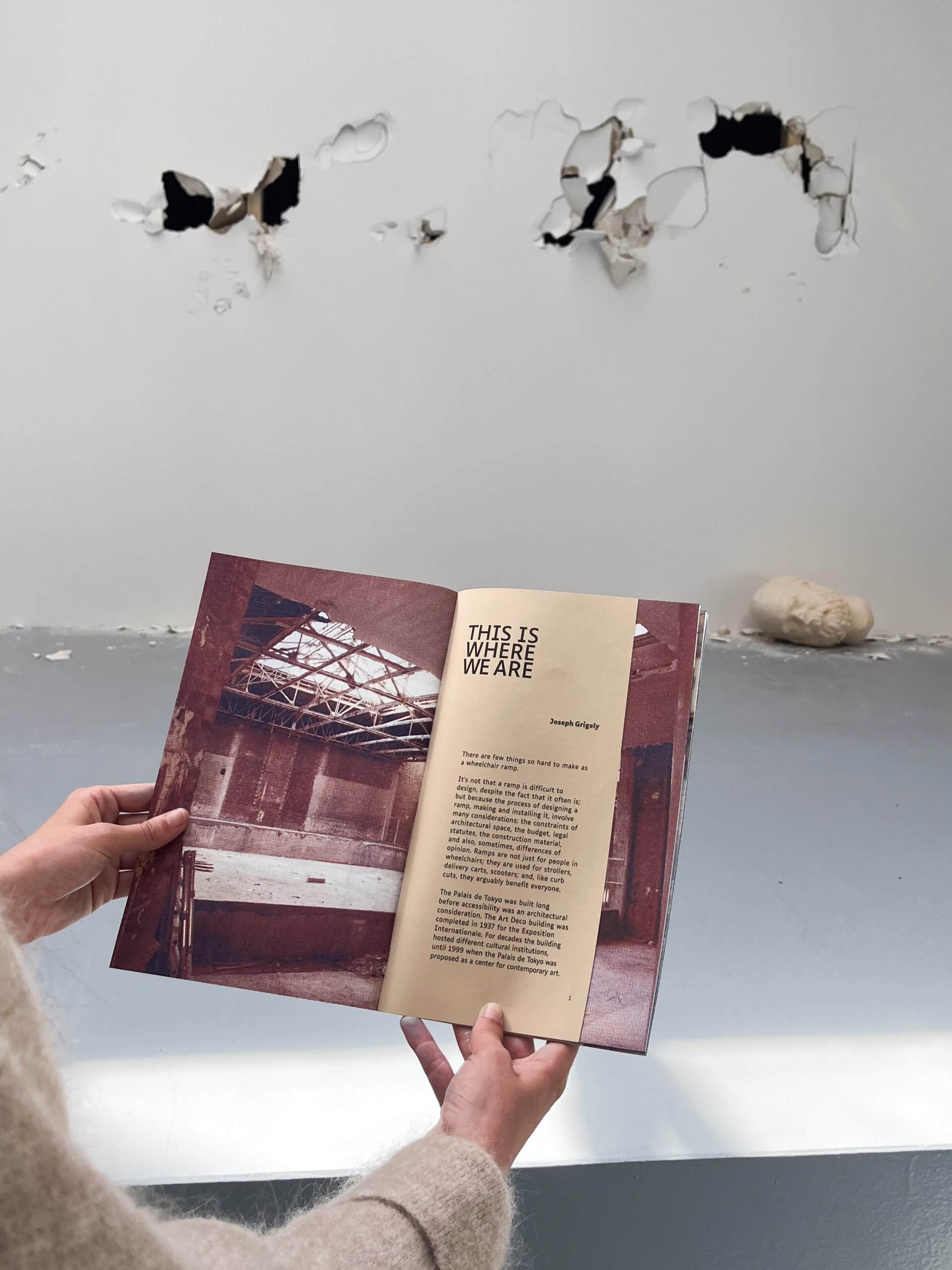 Person holding an open book in front of a white wall with multiple holes and dents; the book shows a photo of a damaged interior and text titled “THIS IS WHERE WE ARE.”.