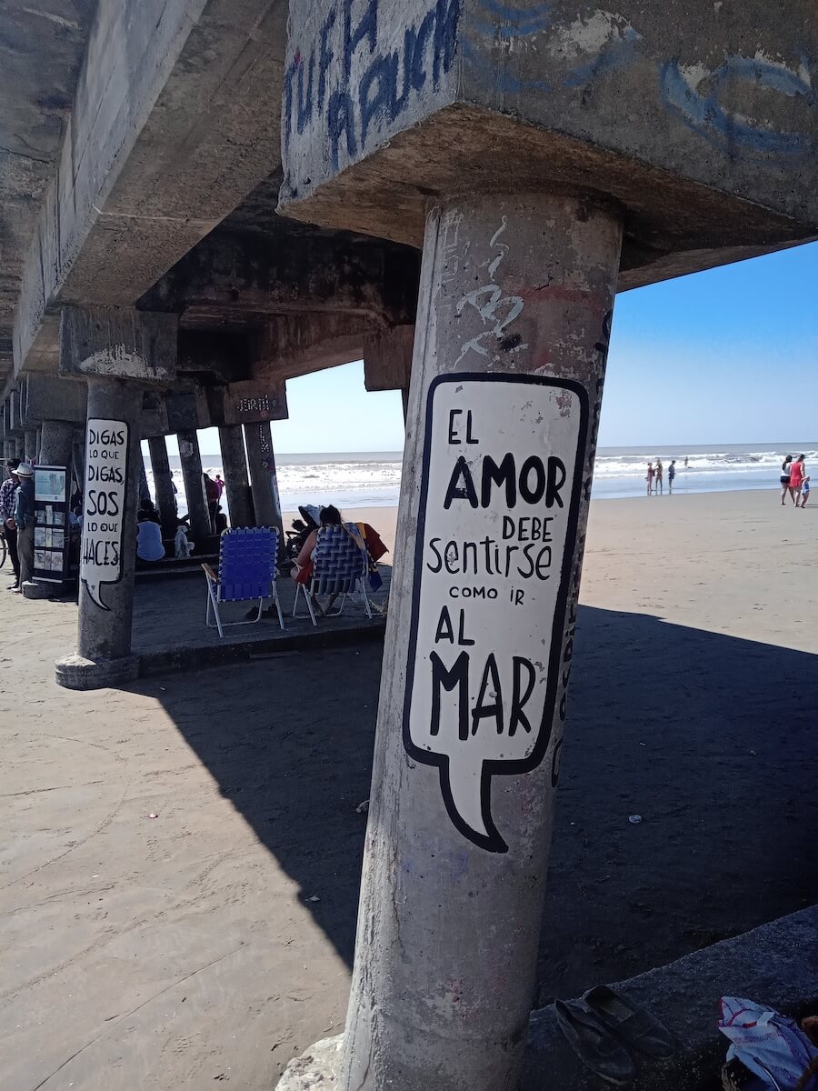 Piliers en béton sous une jetée sur une plage, l'un d'entre eux comportant un graffiti espagnol sur lequel on peut lire El amor debe sentirse como ir al mar. Des personnes et des chaises sont visibles à l'arrière-plan, près du rivage.