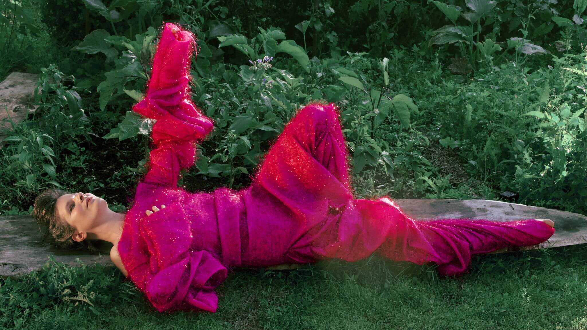 Personne allongée sur une planche de bois en plein air, vêtue d'une tenue texturée magenta vif, entourée de plantes vertes et d'herbe.
