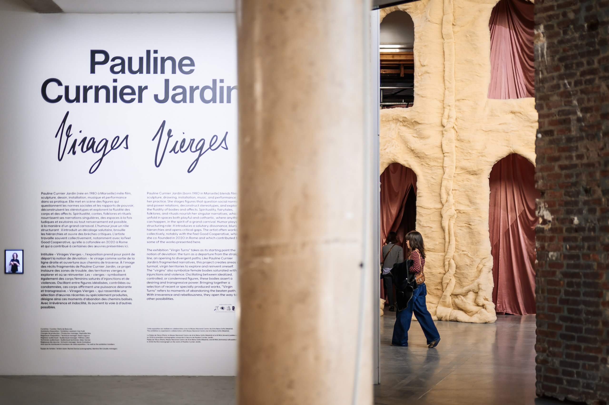 Une femme passe devant un grand texte mural présentant l'exposition Jardins Virages Vierges de Pauline Curnier Jardin, avec des murs sculpturaux beiges et des rideaux roses en arrière-plan.