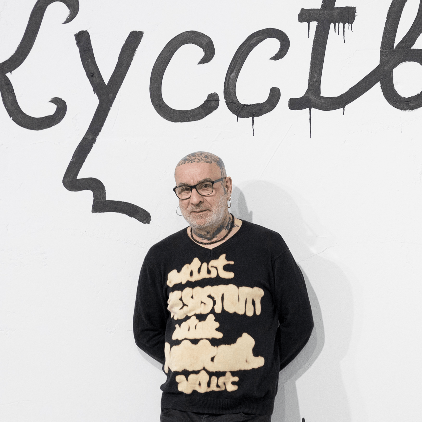 Un homme avec des lunettes et des tatouages se tient devant un mur blanc avec un texte noir de style graffiti, portant un pull noir avec un texte beige.