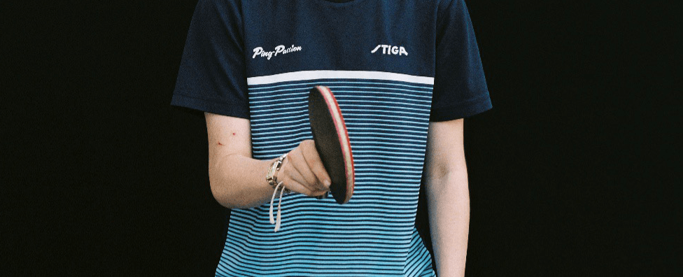 Une personne portant un t-shirt bleu rayé STIGA tient une raquette de tennis de table devant un fond noir.