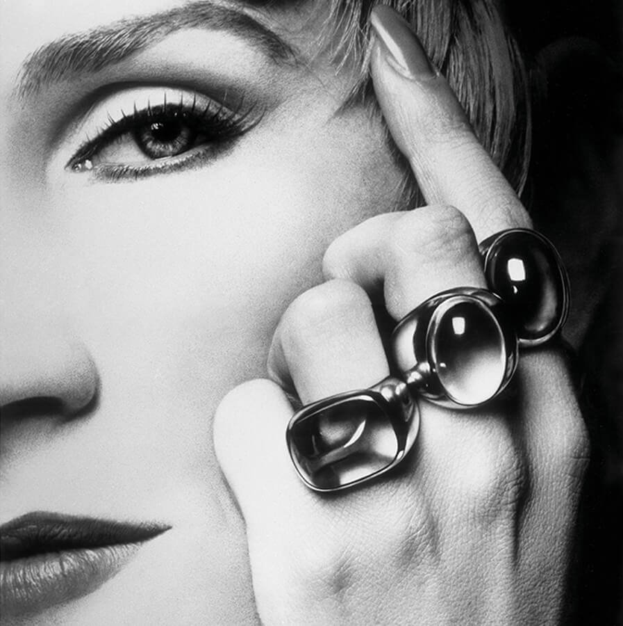 Black and white close-up of a womans face with her hand near her eye, wearing three large, geometric rings on her fingers.