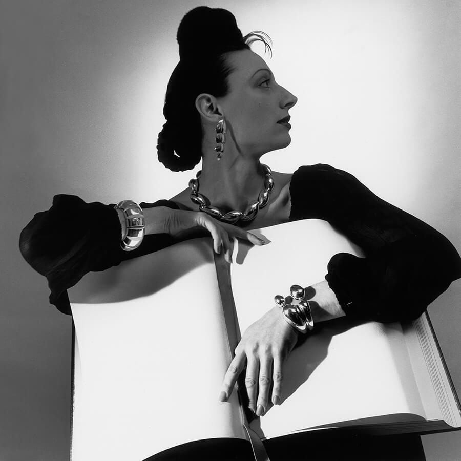 Une femme de profil, portant des bijoux audacieux et des vêtements sombres, repose ses bras sur un grand livre ouvert aux pages vierges, sur un fond uni.