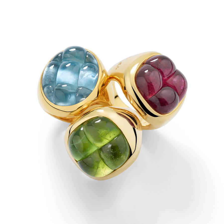 Trois bagues en or ornées de grandes pierres précieuses taillées en cabochon de couleur bleue, verte et rouge, disposées en grappe sur un fond blanc.
