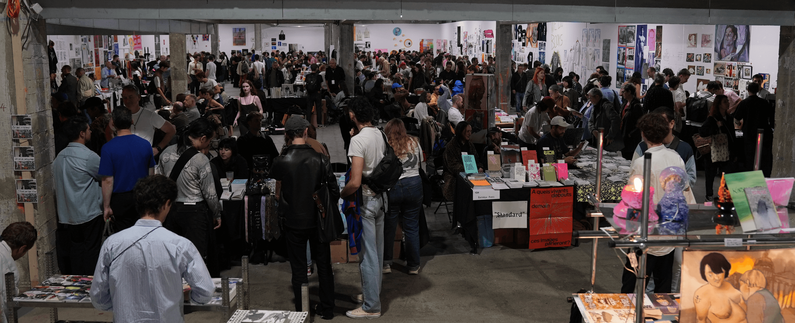 Grande foire d'art intérieure avec des tables bondées présentant des livres, des gravures et des œuvres d'art ; les visiteurs parcourent les expositions et interagissent avec les vendeurs dans un hall spacieux et bien éclairé.