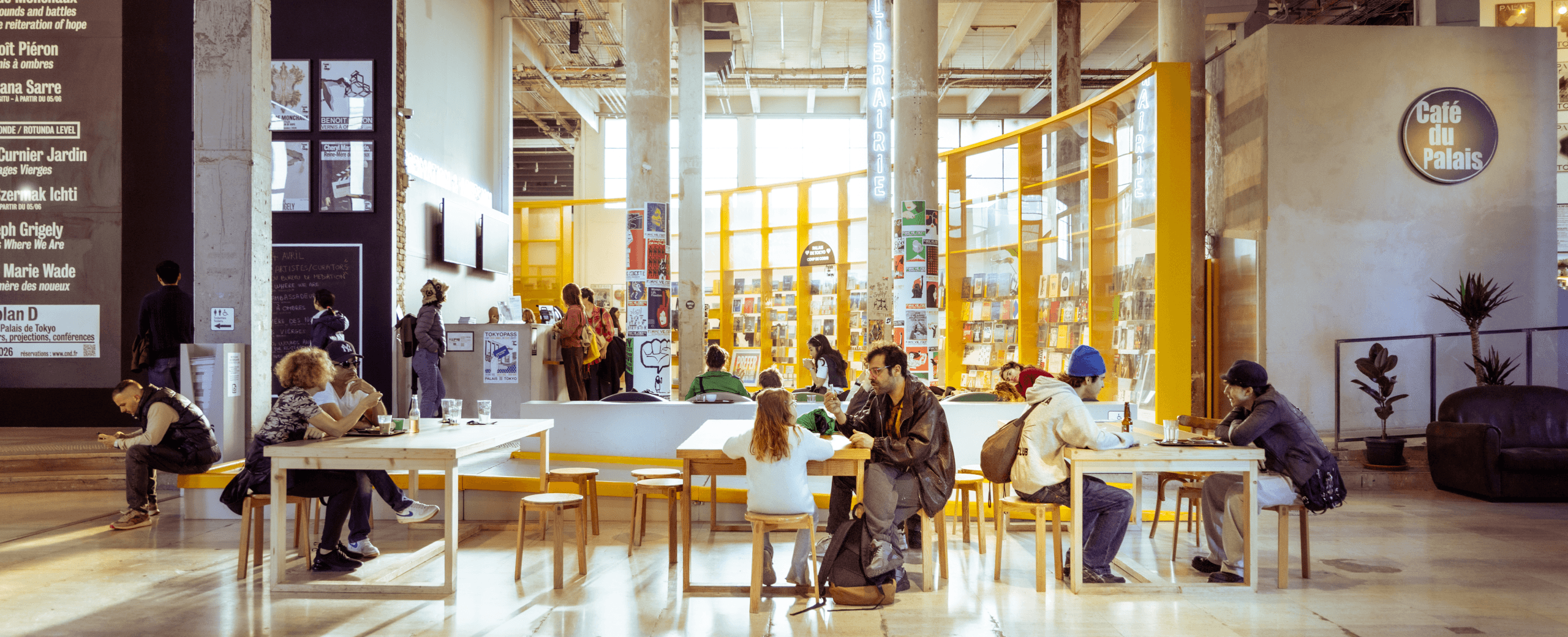 Les gens s'assoient à de longues tables dans un café moderne avec de hauts plafonds, de grandes fenêtres et un mur d'exposition de livres jaune vif à l'arrière-plan.