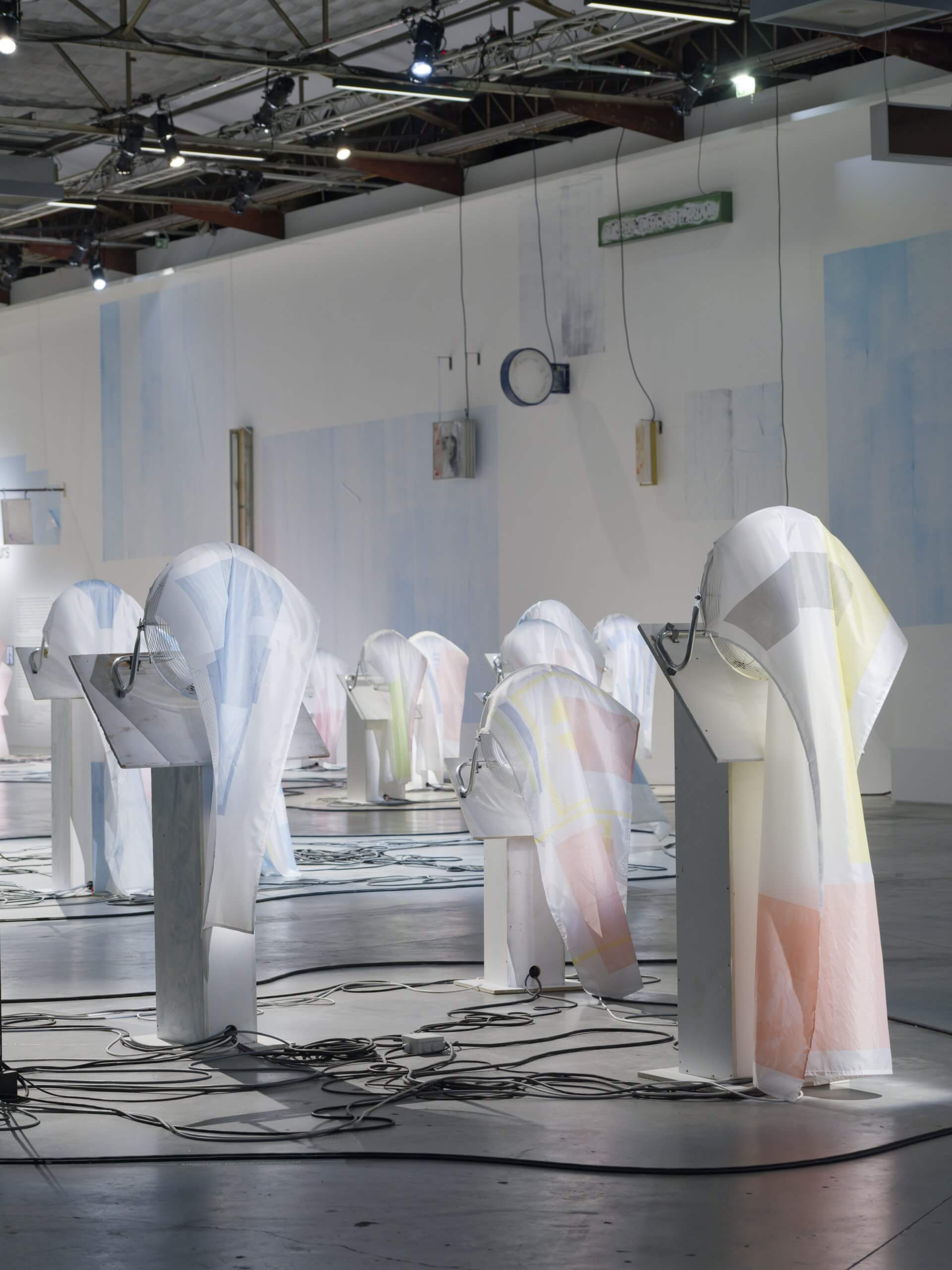 A room with abstract, fabric-draped structures on pedestals, connected by cables, under industrial lighting in a modern exhibition space.