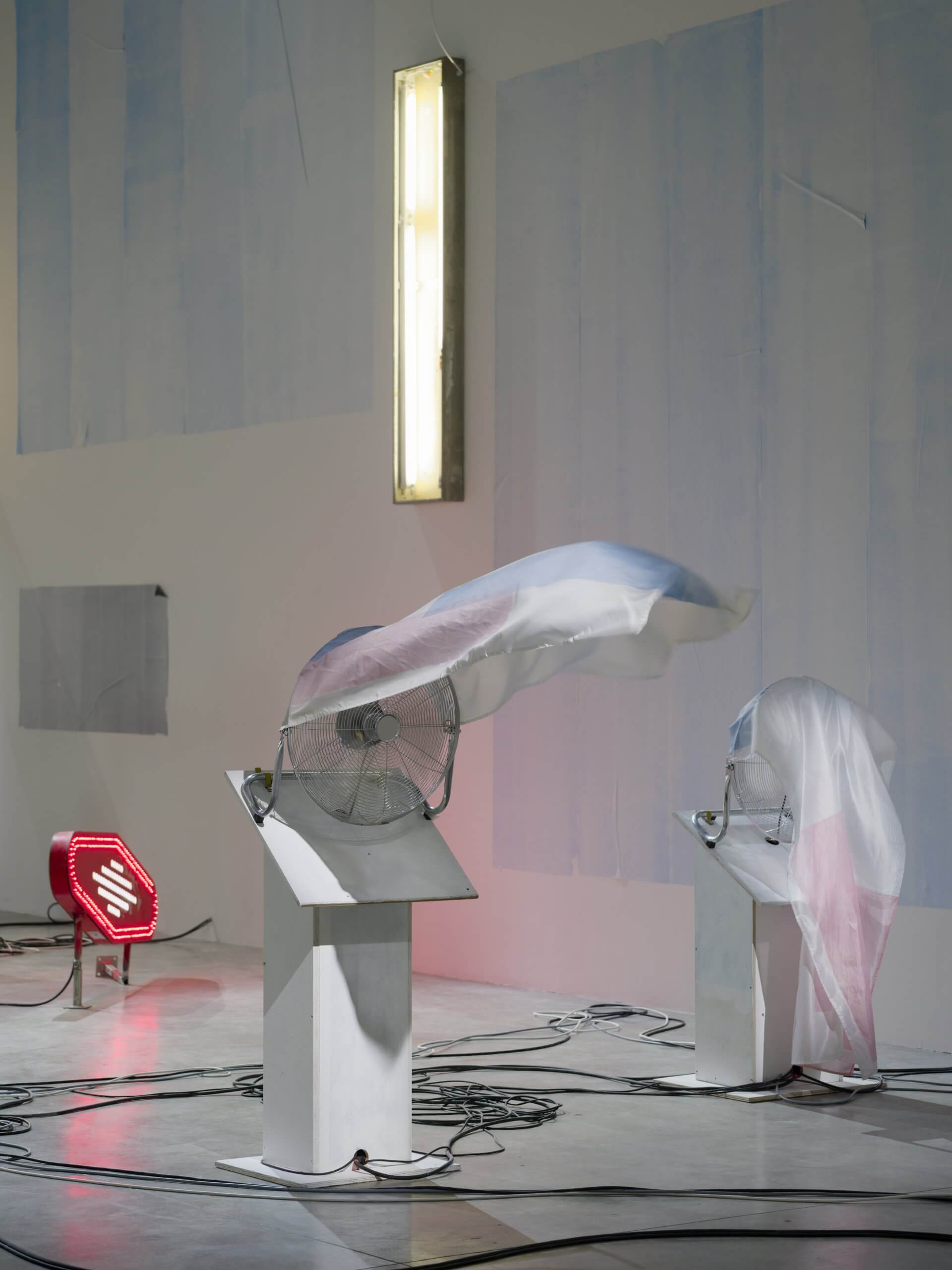 Two pedestal fans covered with sheer plastic sheets blow fabric upwards in an art gallery space, with visible wires on the floor and wall-mounted lights in the background.